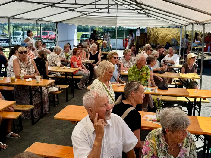 Menschen sitzen und stehen unter einem weißen Zelt an Biertischen und unterhalten sich. Einige halten Getränke in der Hand.