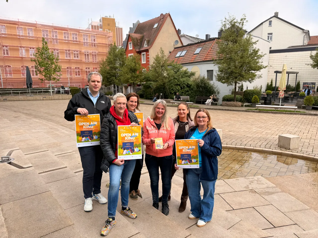 Sechs Personen stehen auf einem Platz vor bunten Fachwerkhäusern und halten Plakate mit der Aufschrift „Open Air Kino“ in den Händen.
