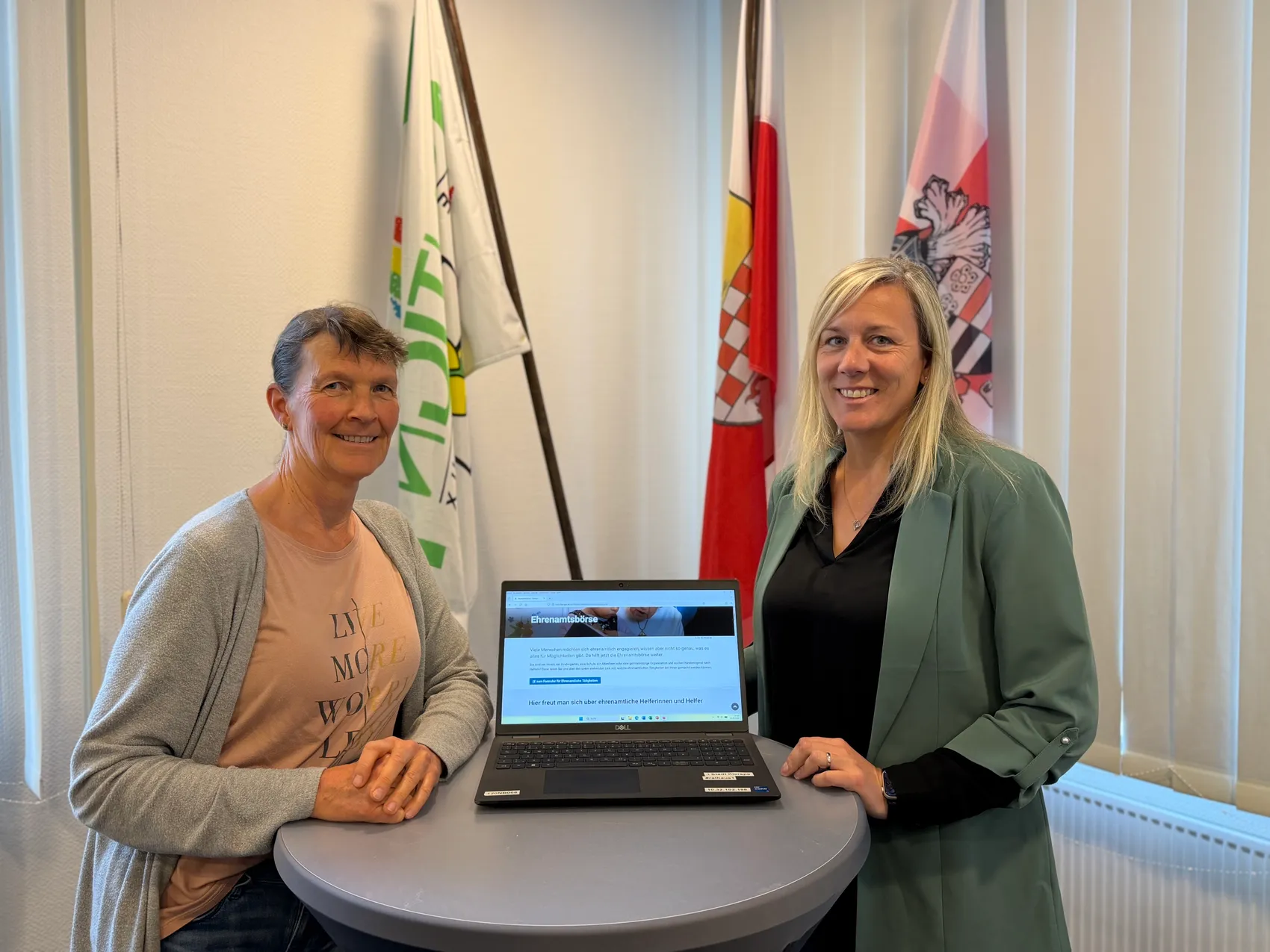 Zwei Frauen sitzen nebeneinander vor einem Laptop mit der geöffneten Ehrenamtsbörse auf dem Bildschirm. Im Hintergrund hängen Flaggen.