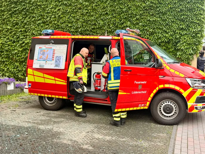 Zwei Feuerwehrleute in Schutzkleidung stehen vor einem roten Mannschaftswagen der Feuerwehr Lüdenscheid, der mit technischer Ausrüstung bestückt ist. Im Hintergrund ist dichter grüner Buschwerk zu sehen.