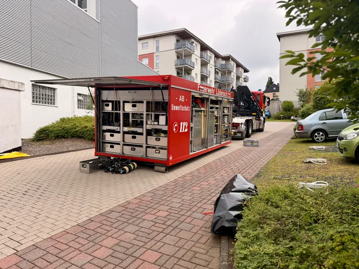 Roter Mannschaftswagen der Feuerwehr Lüdenscheid mit geöffneten Türen und Ausrüstungsteilen davor, im Hintergrund ein Wohngebäude und ein parkendes Auto. Der Schriftzug 