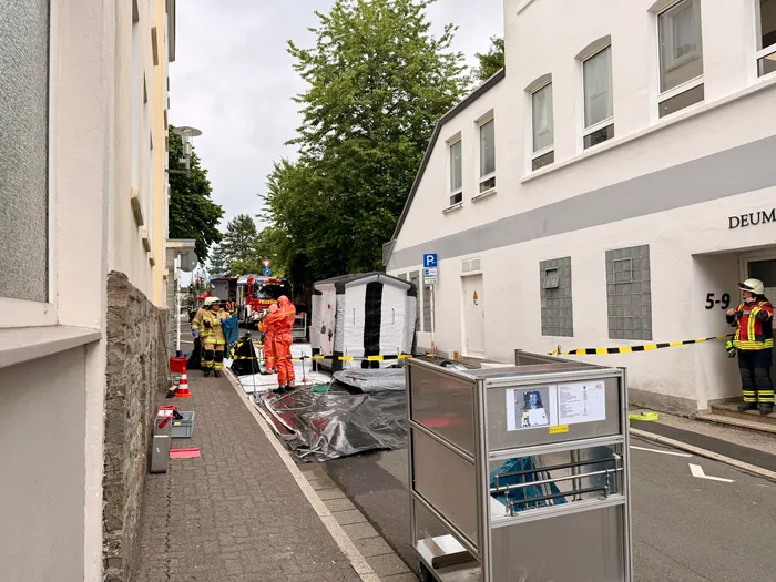 Feuerwehrleute in Schutzanzügen sichern einen Bereich vor einem Mehrfamilienhaus mit Absperrband. Im Hintergrund steht ein aufblasbarer Zelt.