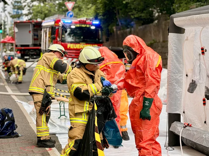 Feuerwehrleute in Schutzkleidung helfen sich gegenseitig beim Anlegen von Atemschutzmasken und Chemieschutzanzügen nach einem Chlorgasunfall. Im Hintergrund steht ein rotes Feuerwehrauto.