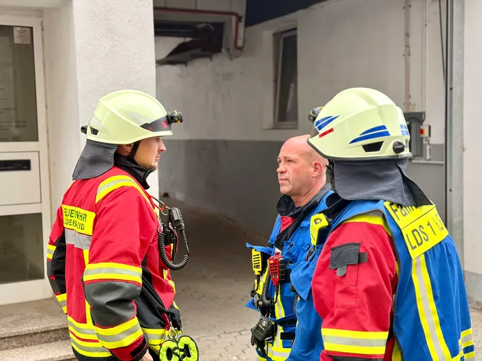 Drei Feuerwehrleute mit Schutzhelmen und roter Einsatzkleidung besprechen die Lage vor einem Gebäude mit roten Fassadenflächen. Einer der Feuerwehrleute trägt eine Atemmaske.