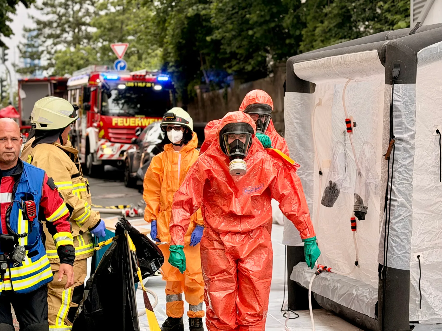 Feuerwehrleute in Schutzanzügen und Atemschutzmasken betreten einen aufblasbaren Dekontaminationszelt während eines Chlorgasunfalls in Lüdenscheid. Im Hintergrund ist ein rotes Feuerwehrauto zu sehen.