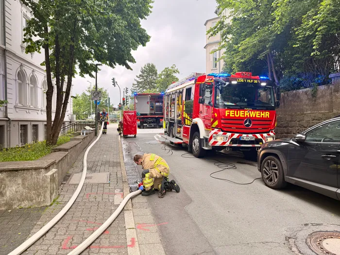 Feuerwehrfahrzeug mit Blaulicht steht an einem Bürgersteig, während ein Feuerwehrbeamter an einem Schlauch arbeitet. Im Hintergrund sind Gebäude und Bäume zu sehen.