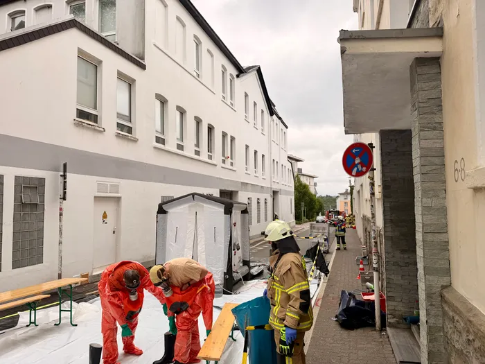 Mehrere Einsatzkräfte in Schutzanzügen und ein Feuerwehrmann stehen vor einem Gebäude, das von einem Chlorgasunfall betroffen ist. Im Hintergrund ist ein Zelt und weitere Einsatzkräfte zu sehen.