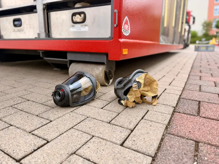 Zwei Atemschutzmasken liegen auf einem Pflaster vor einem roten Anhänger mit Aufschrift „Feuerwehr Lüdenscheid“. Im Hintergrund ist ein Wohngebäude zu sehen.