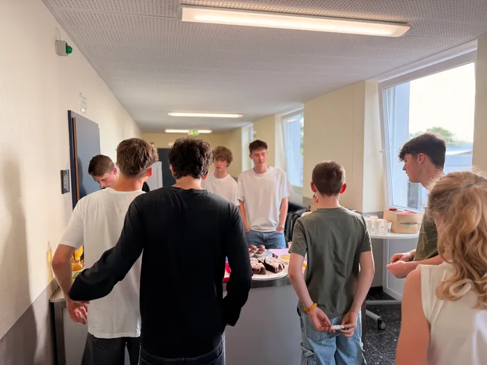 Eine Gruppe Jugendlicher steht in einem hellen Flur um einen Tisch mit Snacks und Getränken. Sie unterhalten sich und scheinen eine Veranstaltung zu genießen.