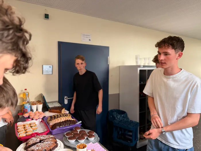 Junge Leute stehen um einen Tisch mit Kuchen und Gebäck. Im Hintergrund ist eine offene Tür zu sehen, durch die ein weiterer junger Mann blickt.