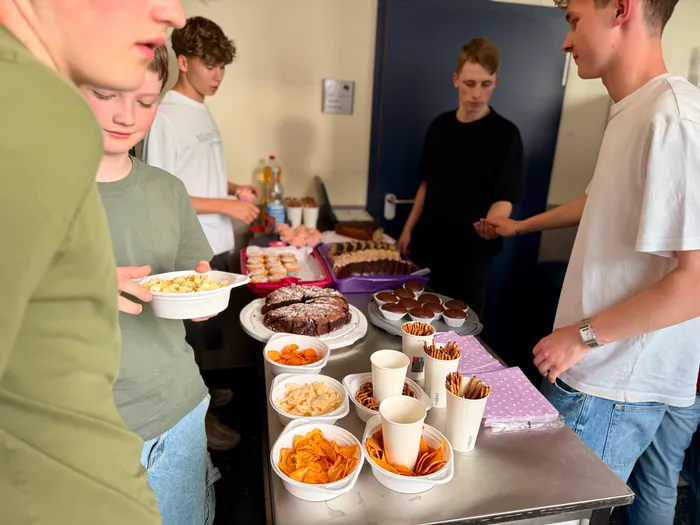 Junge Leute stehen um einen Tisch mit einer großen Auswahl an Snacks und Getränken. Im Hintergrund weitere Besucher des Bunten Abends der Abiturienten.