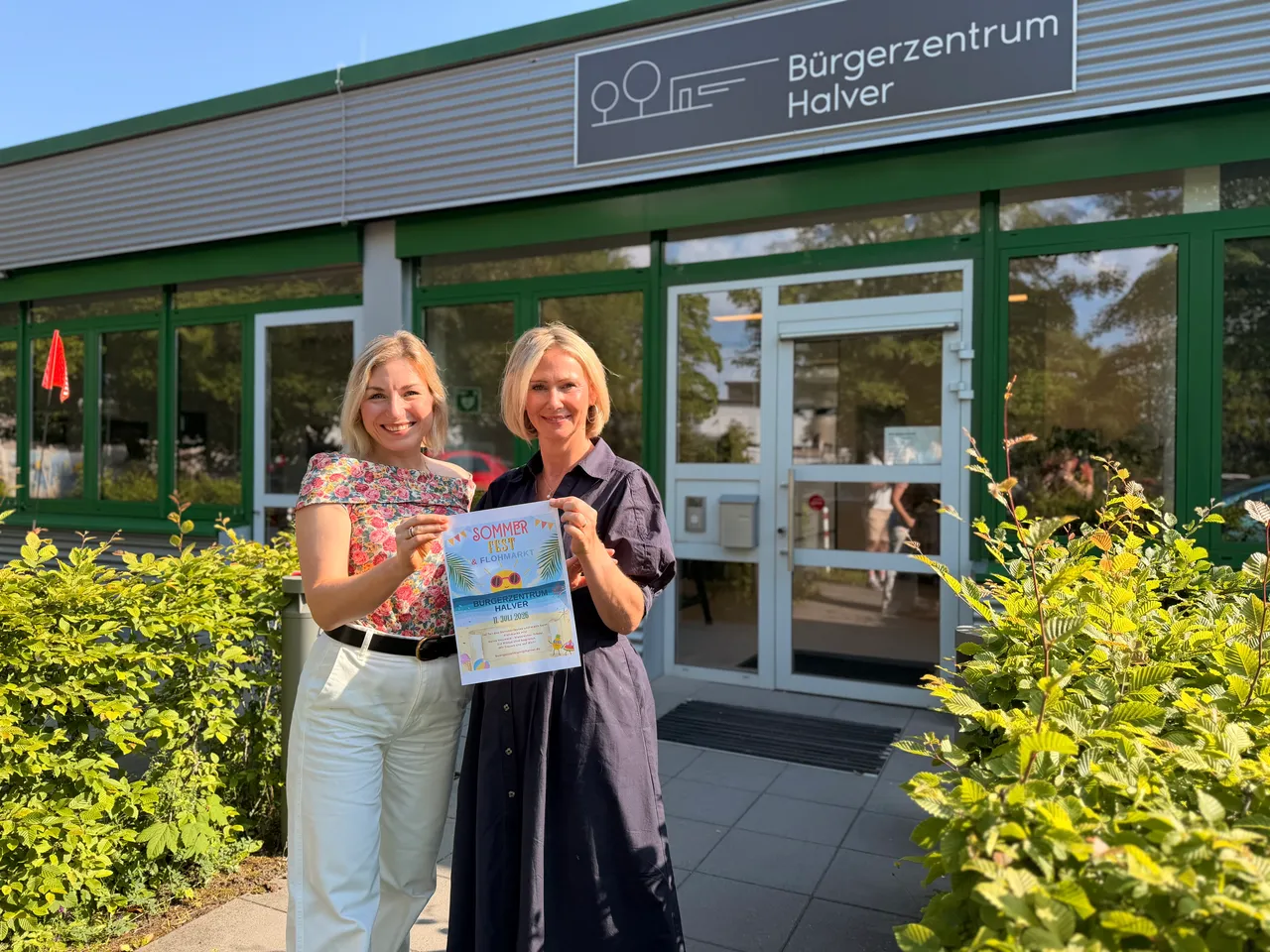 Zwei Frauen halten vor dem Bürgerzentrum Halver ein farbiges Plakat für ein Sommerfest hoch. Im Hintergrund ist die gläserne Eingangstür des Bürgerzentrums zu sehen.