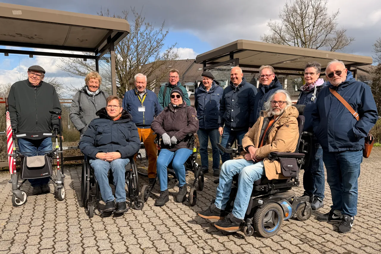 Gruppe von etwa zehn Personen unterschiedlichen Alters posiert im Freien vor einer modernen Bushaltestelle. Einige nutzen Rollstühle oder Gehhilfen.