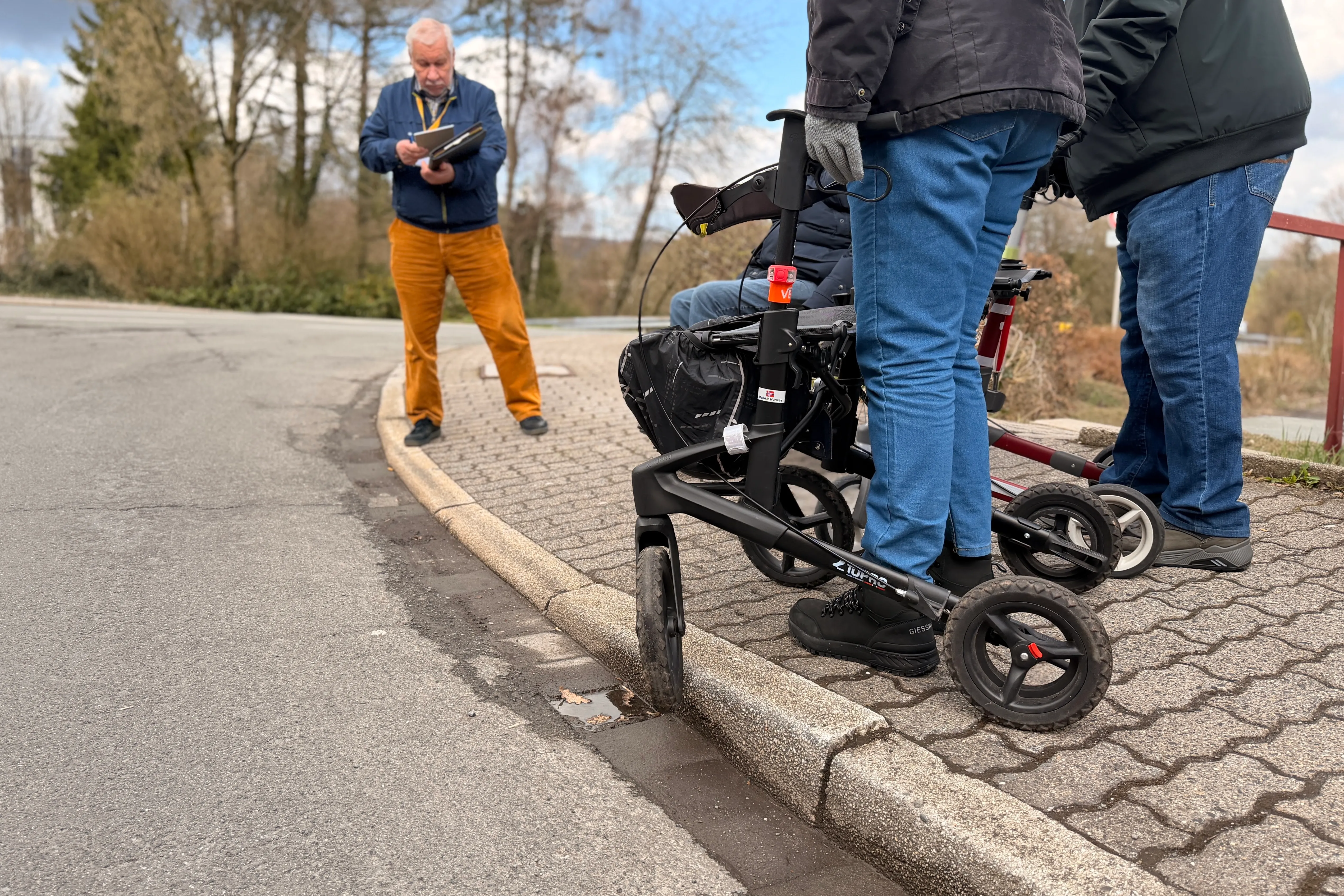 Eine Person mit Gehbehinderung nutzt einen Rollator auf einem Bürgersteig, während ein Mann im Hintergrund Notizen macht.