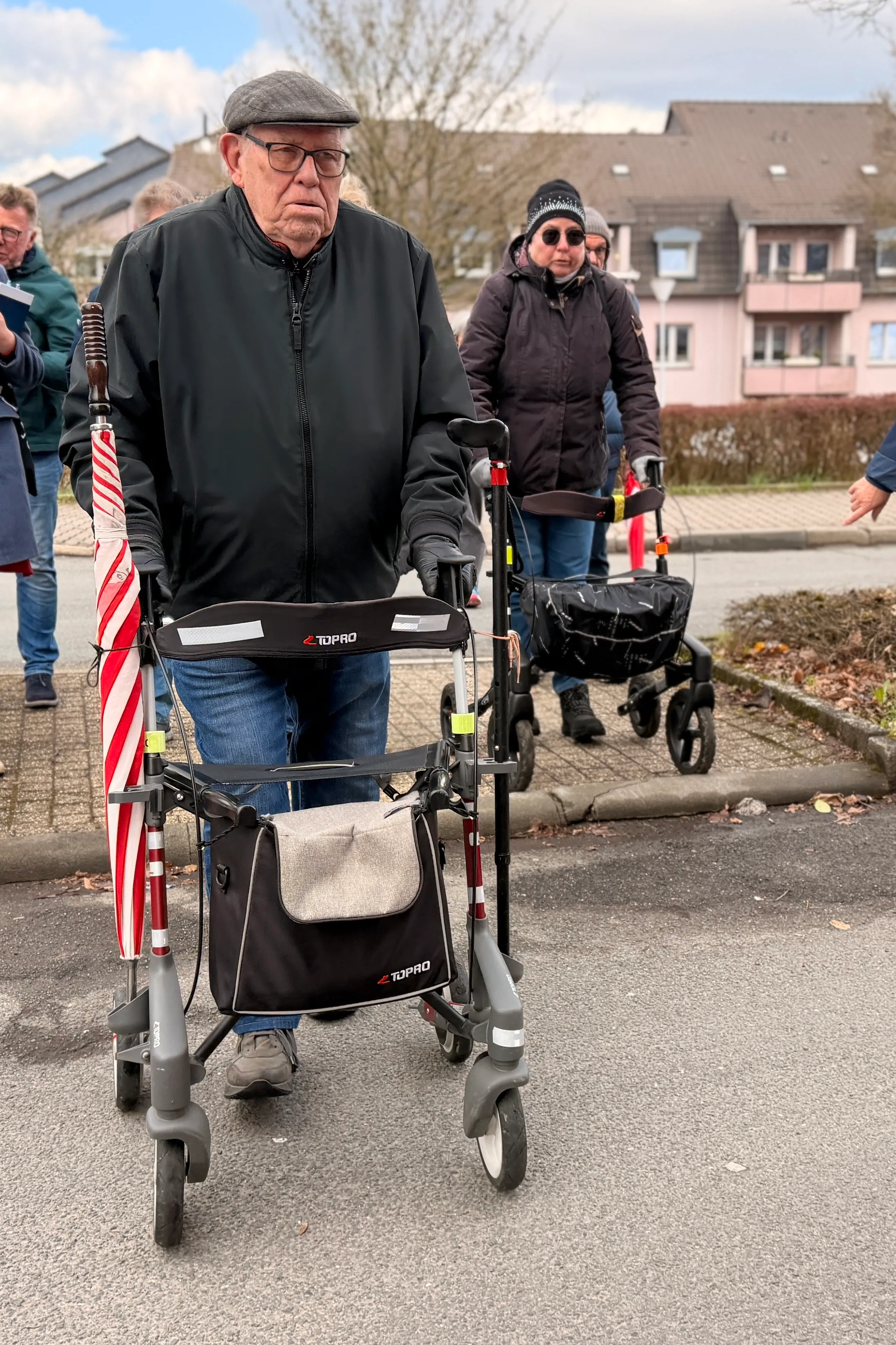 Ein älterer Mann mit Schiebermütze und Rollator schiebt diesen auf einem Bürgersteig vorwärts. Im Hintergrund eine Frau mit Sonnenbrille und Rollator sowie weitere Personen.
