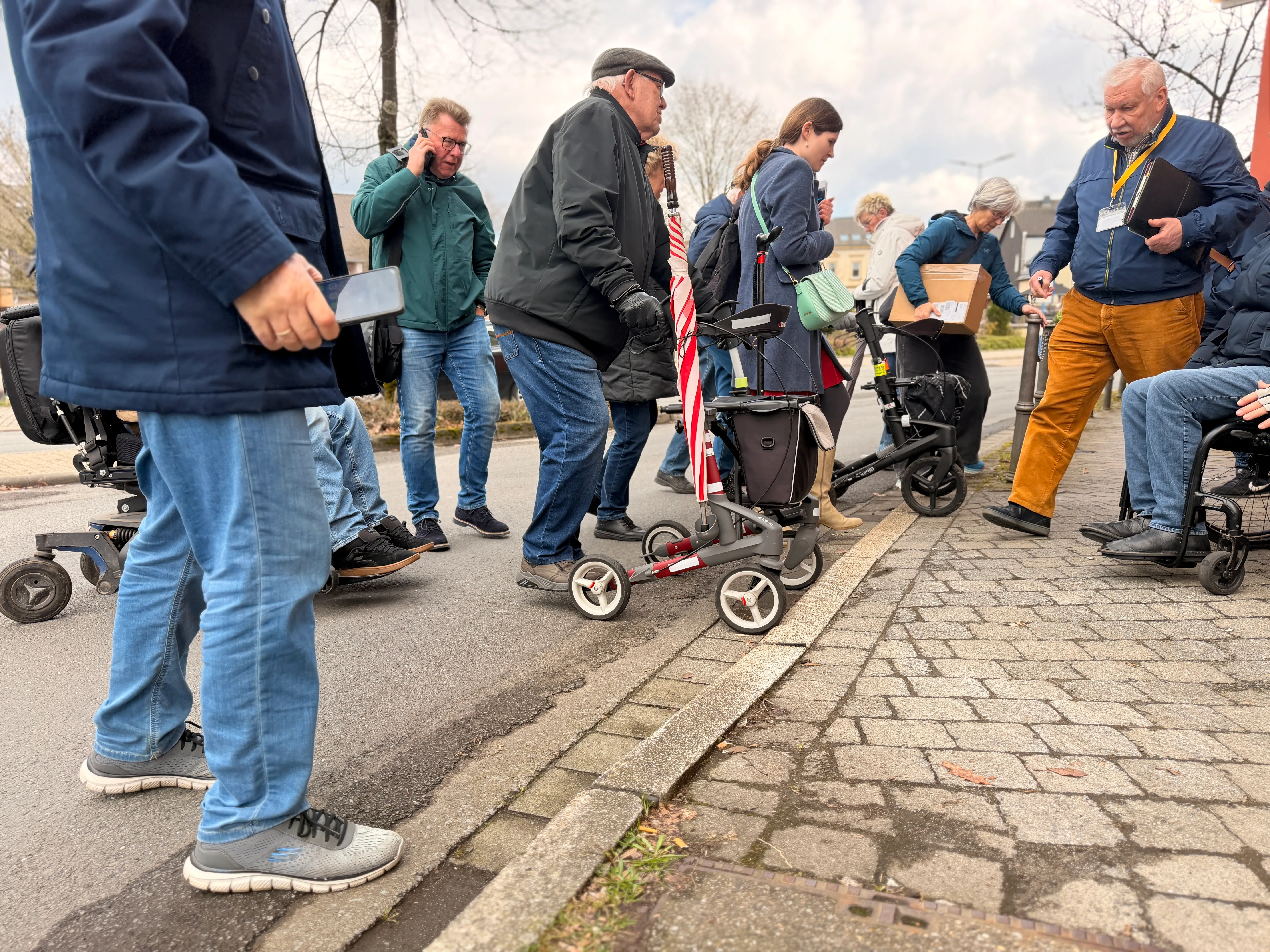 Eine Gruppe von Menschen steht an einer abgesenkten Bordsteinkante, einige mit Rollatoren oder im Rollstuhl. Sie scheinen sich zu unterhalten, während ein Mann am Telefon spricht.
