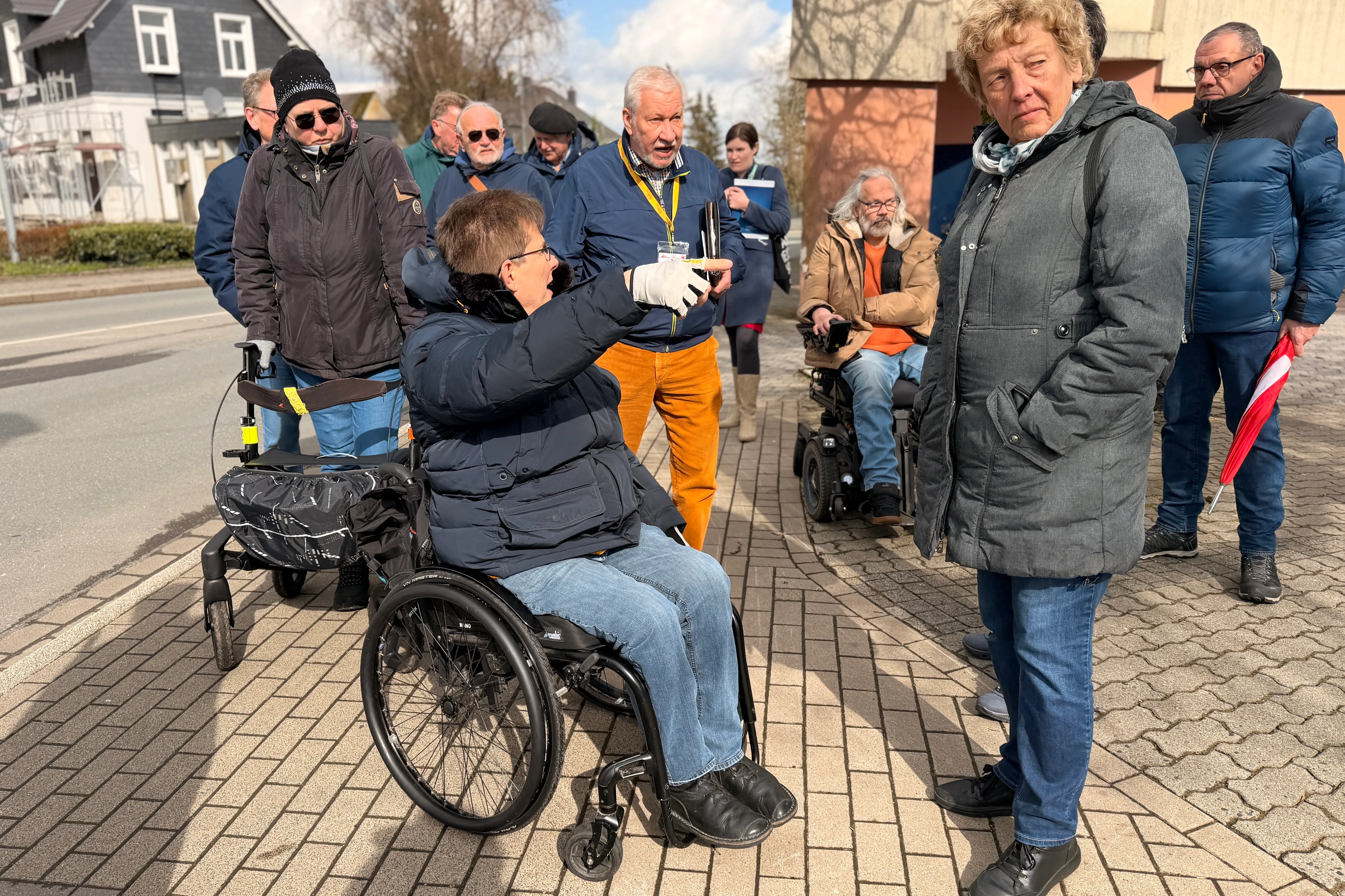 Eine Frau im Rollstuhl und eine Gruppe von Menschen stehen auf einem Gehweg und betrachten eine Barriere. Ein Mann mit einer gelben Weste deutet auf den Gehweg.