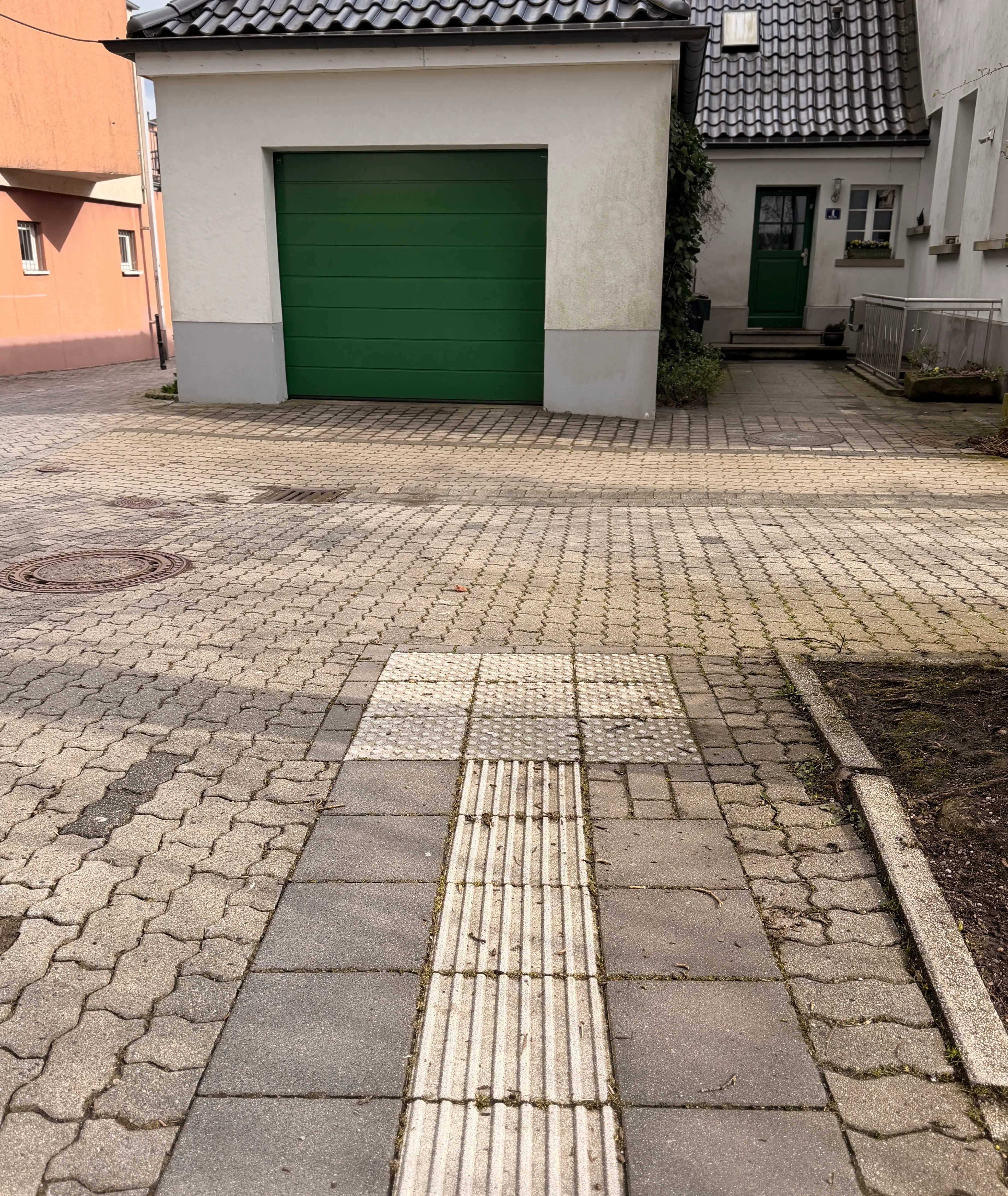 Ein Gehweg mit taktilen Leitsystemen für sehbehinderte Menschen führt zu einer Garage mit grünem Tor und einem Hauseingang.