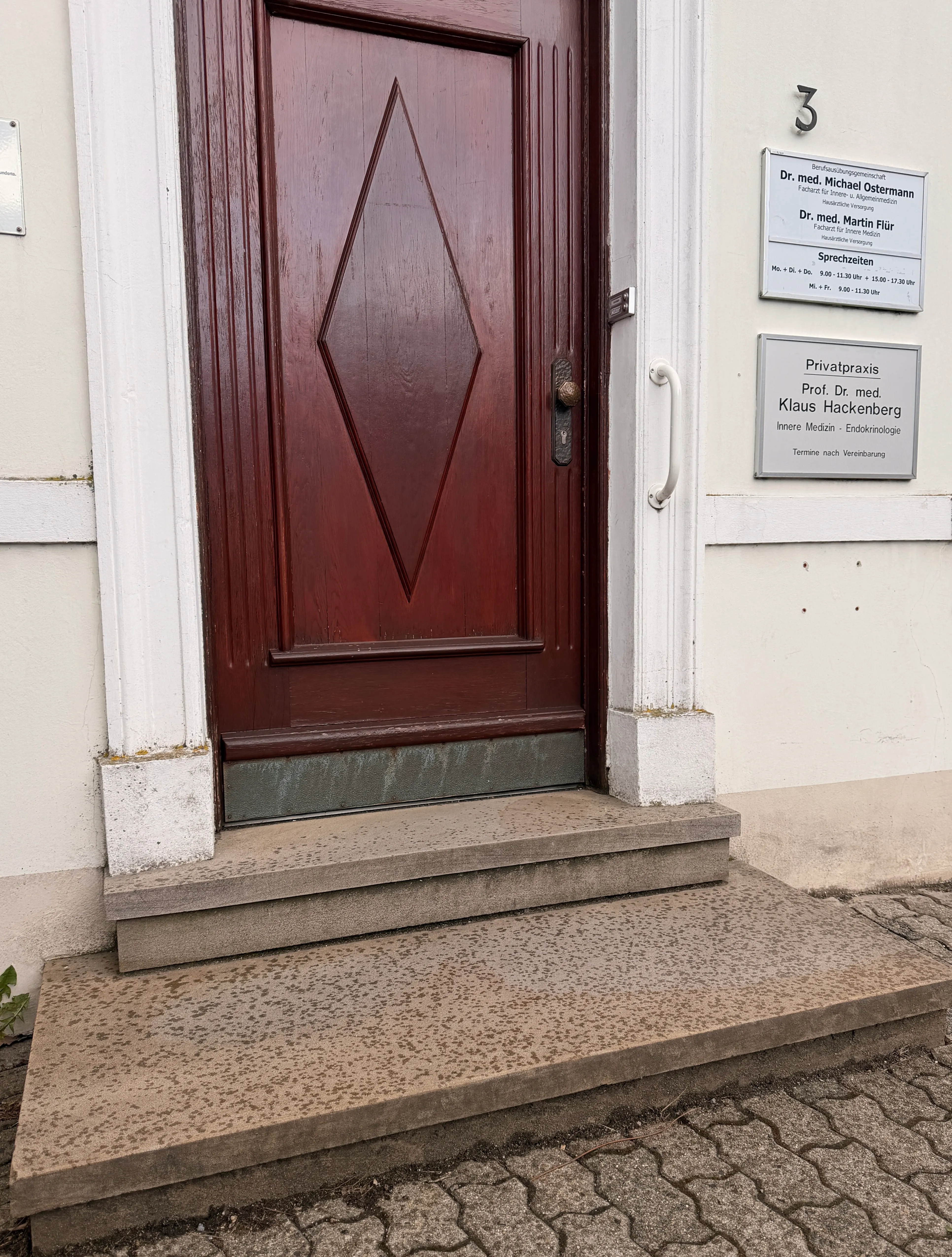 Roter Holzeingang mit Raute-Ornament und Messingbeschlag, daneben ein Klinkersockel und zwei Stufen zur Tür.