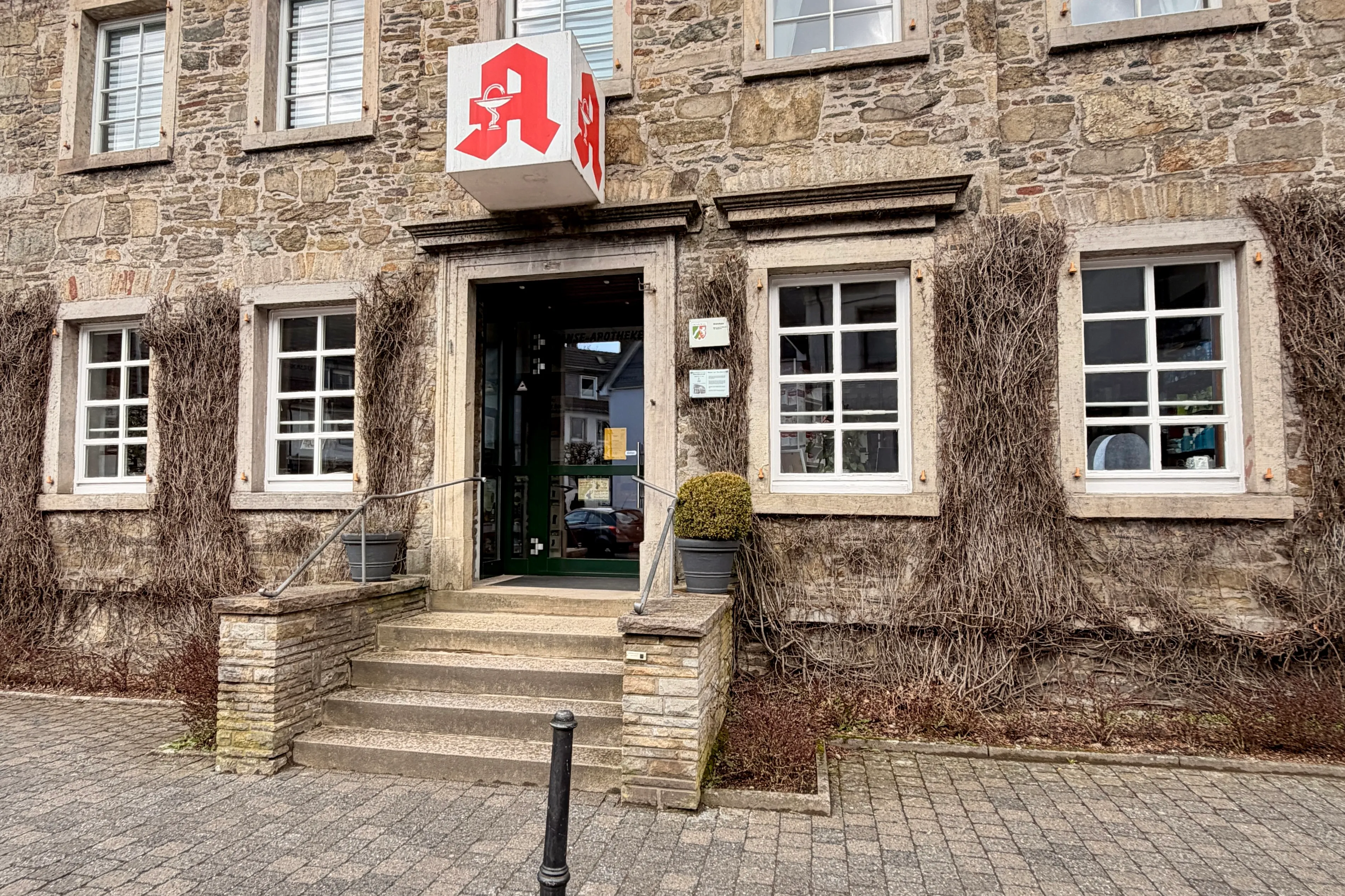 Eingang einer Apotheke mit mehreren Stufen, gekennzeichnet durch ein rotes Schild mit einem weißen Mörser.