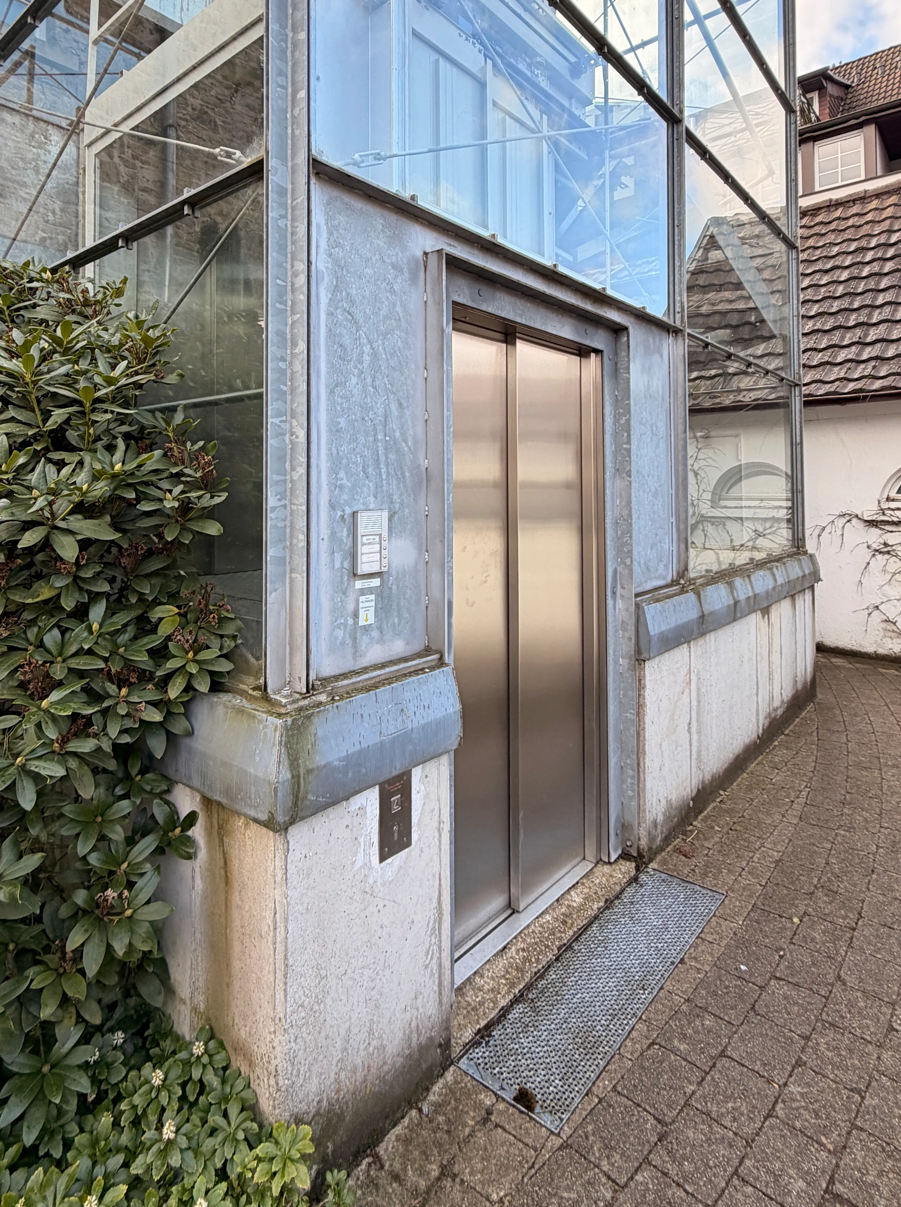 Ein gläserner Fahrstuhl neben dem Hintereingang einer Apotheke mit Kletterpflanzen an der Fassade.
