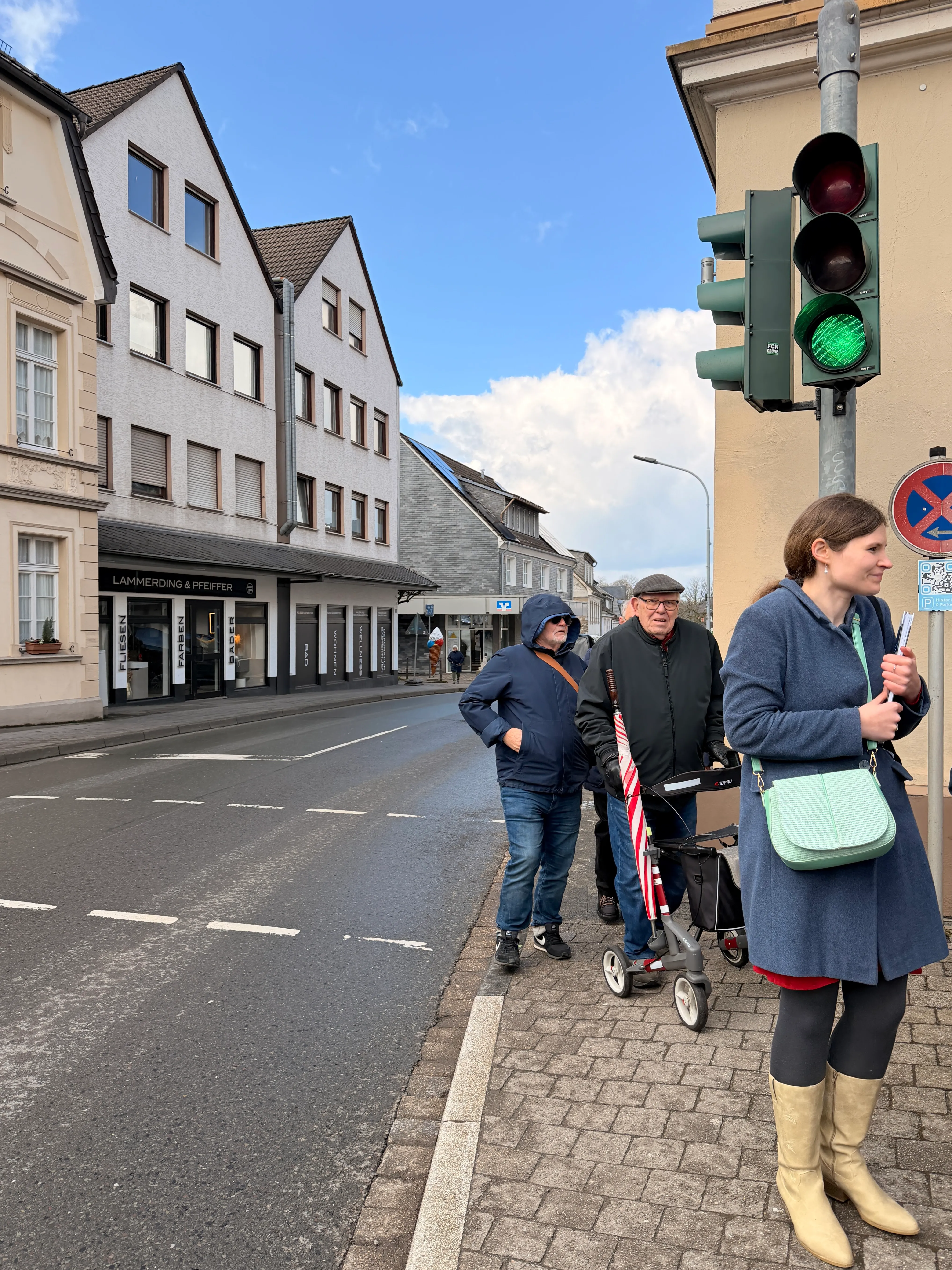 Personen warten an einer schmalen Fußgängerampel vor Fachgeschäften. Ein Rollator steht auf dem Bürgersteig, der sich verengt.