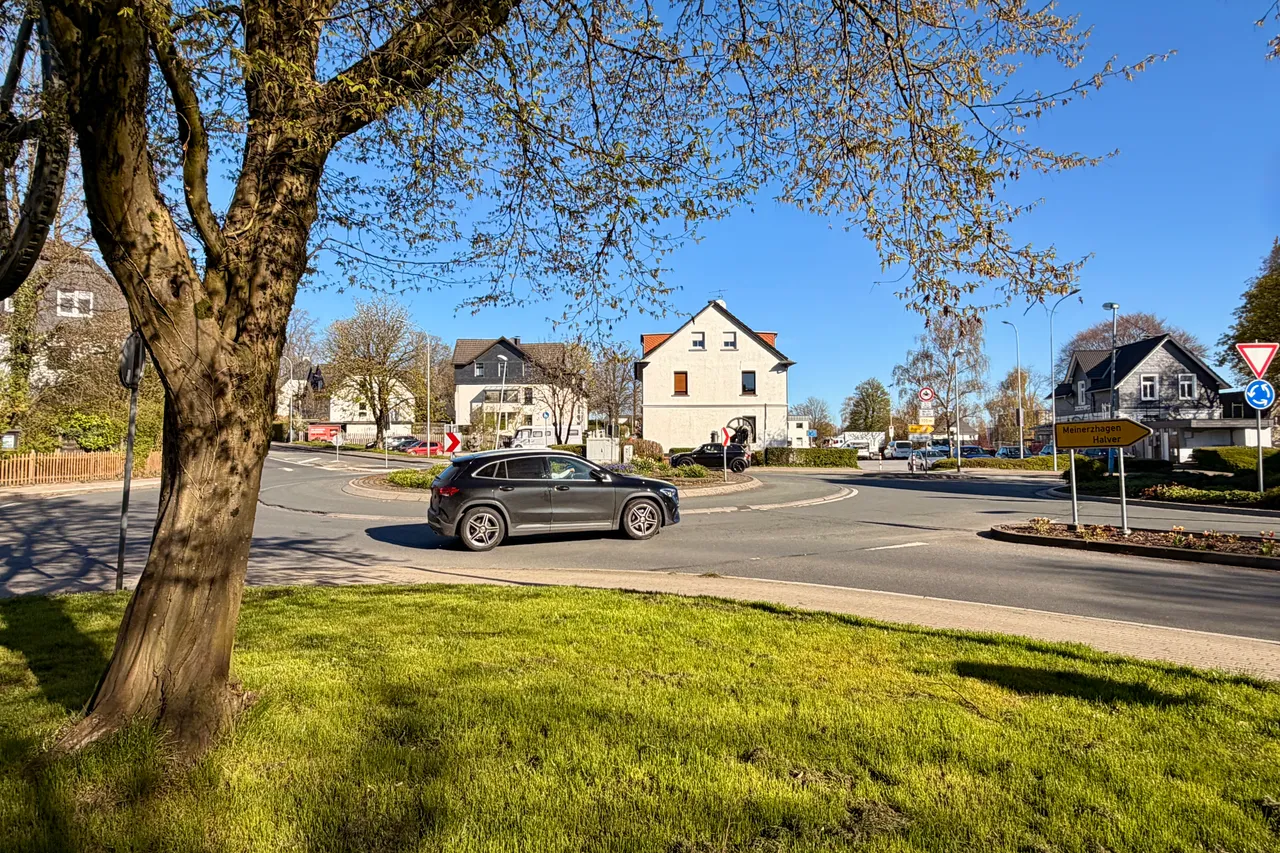 Ein Kreisverkehr mit einem grauen SUV im Verkehr, umgeben von Häusern und Verkehrszeichen an einem sonnigen Tag.