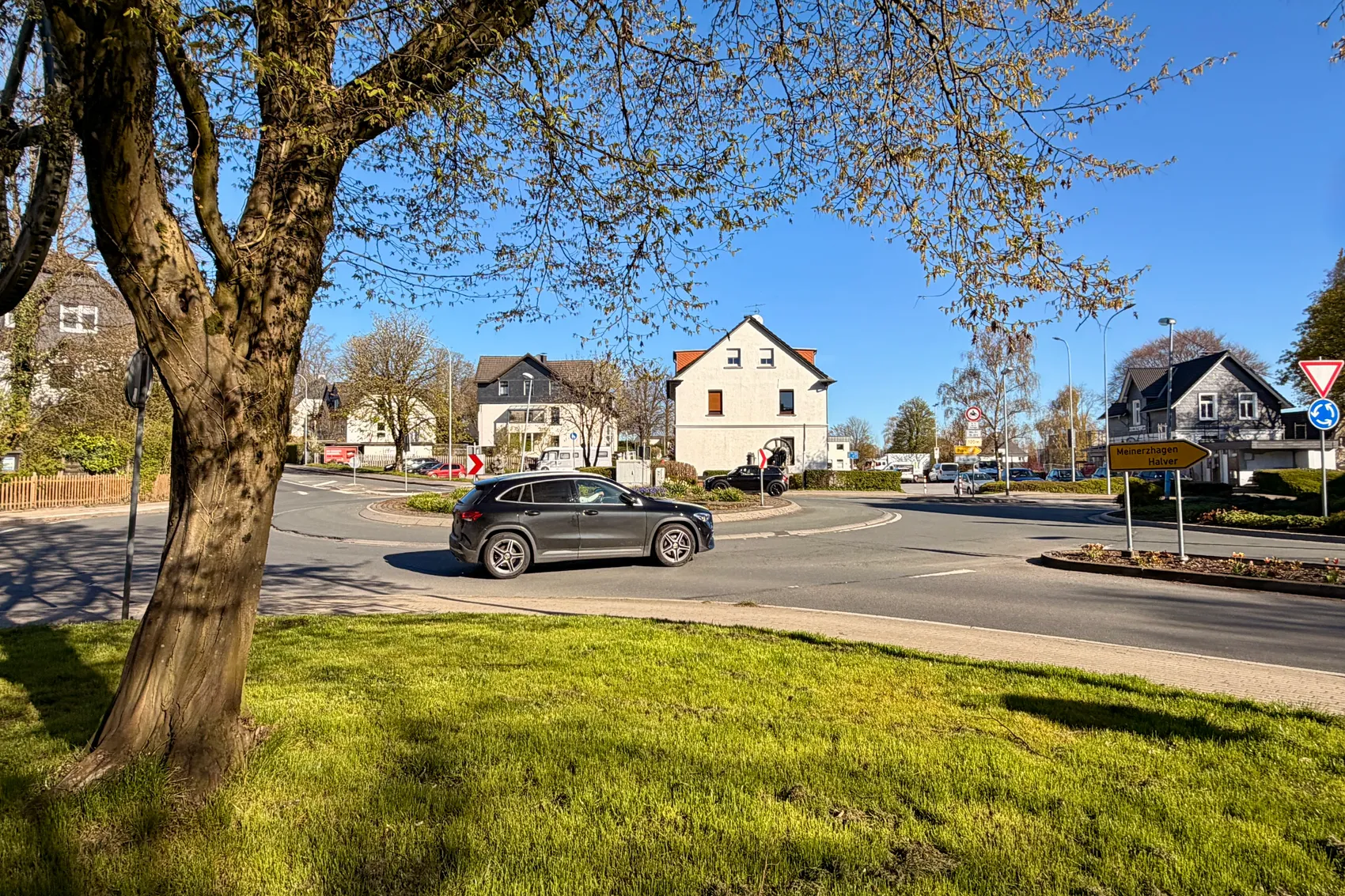 Ein Kreisverkehr mit einem grauen SUV im Verkehr, umgeben von Häusern und Verkehrszeichen an einem sonnigen Tag.