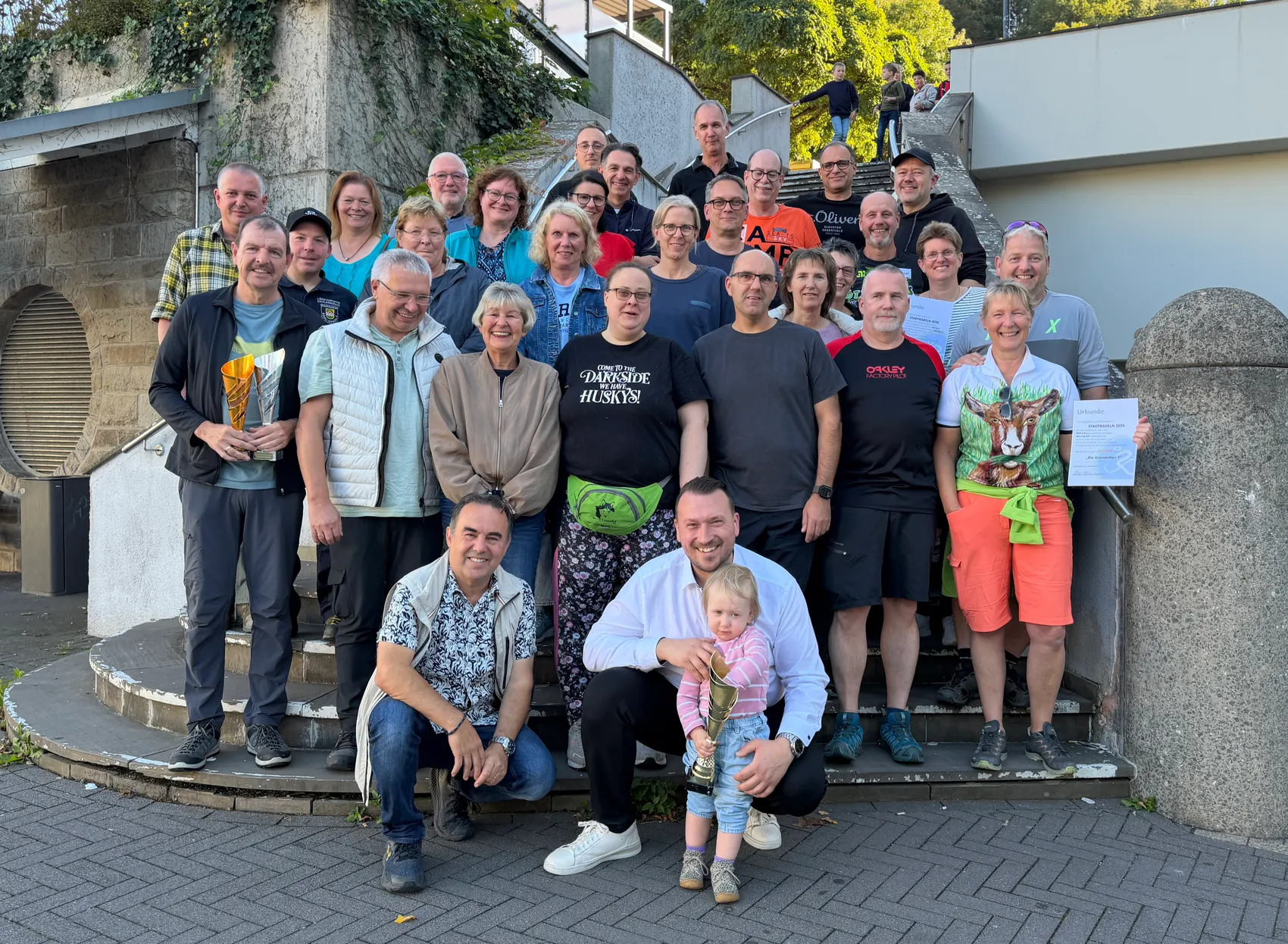 Eine Gruppe von Menschen posiert auf einer Treppe und hält Trophäen sowie eine Urkunde.