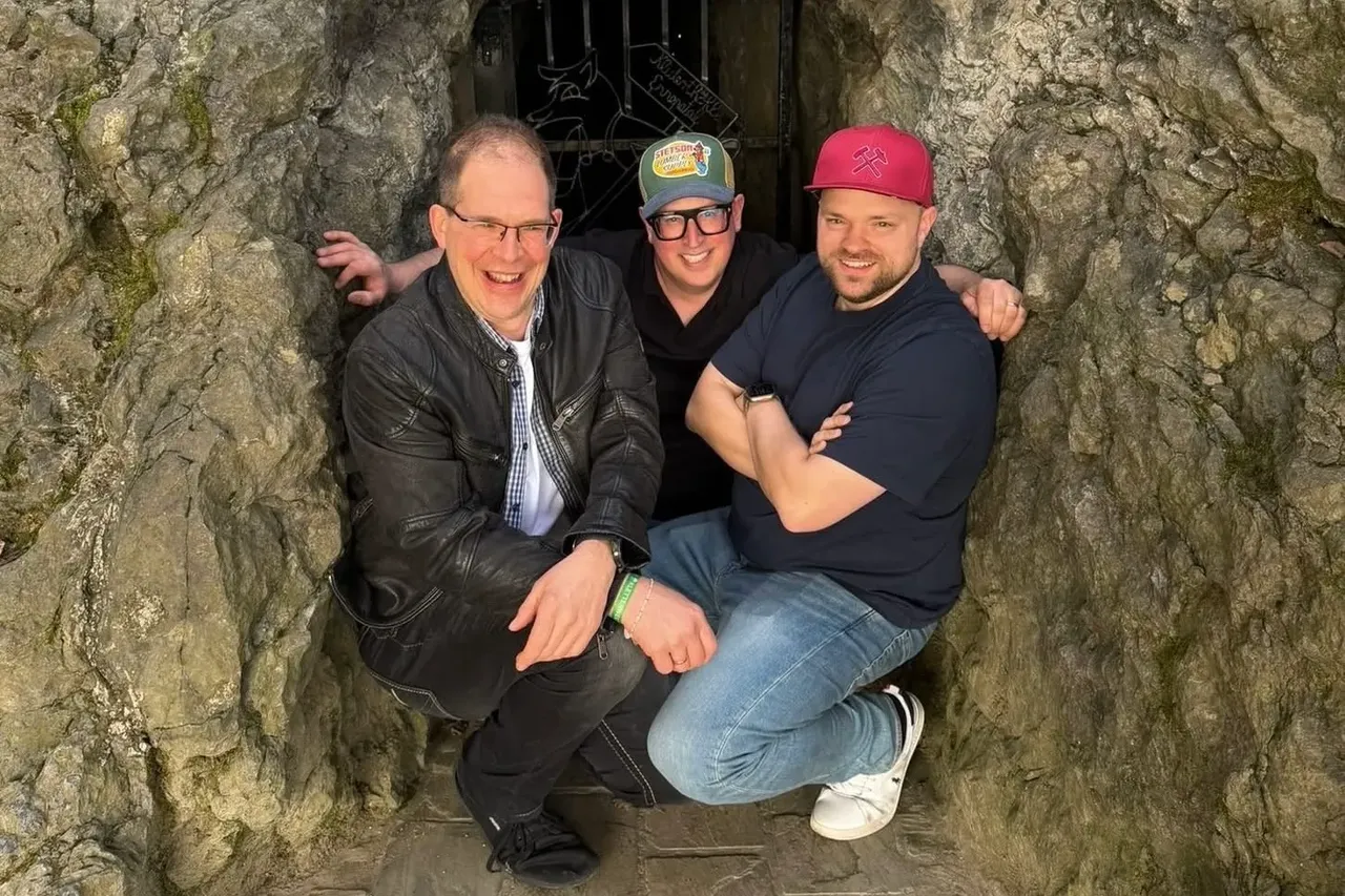 Drei Männer posieren vor einem dunklen Eingang in einer steinernen Wand. Der Mann links trägt eine Lederjacke und Brille, der in der Mitte eine Kappe und der rechts eine Baseballkappe.