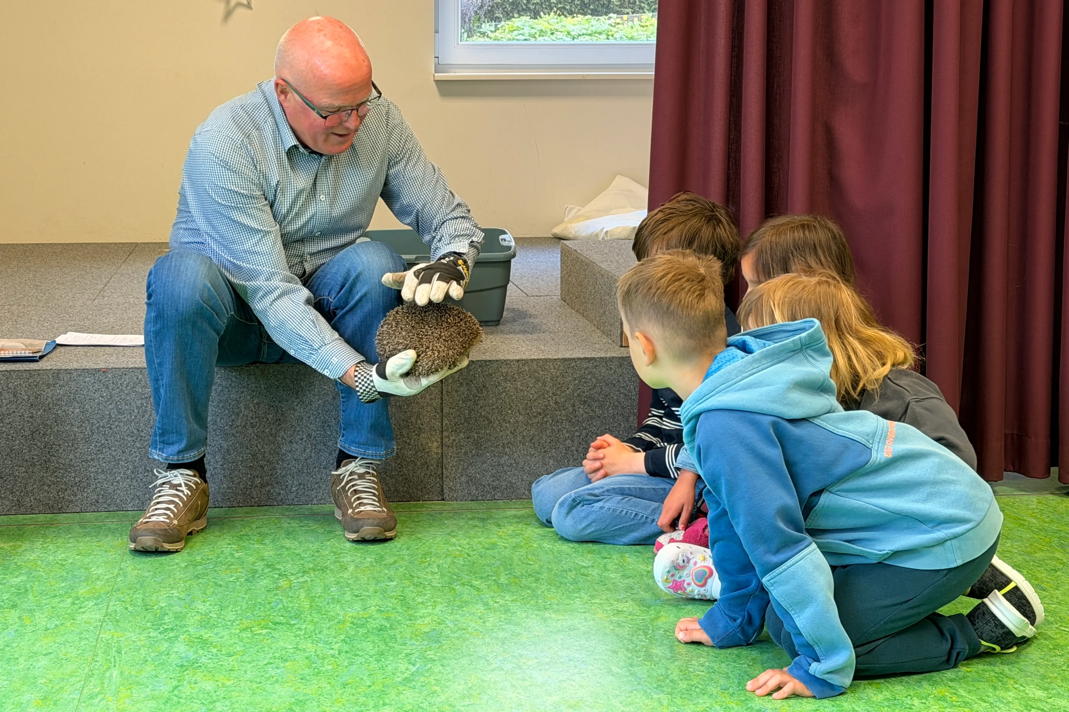 Ein Mann zeigt Kindern einen Igel. Sie sitzen auf dem Boden und schauen dem Tier interessiert zu.
