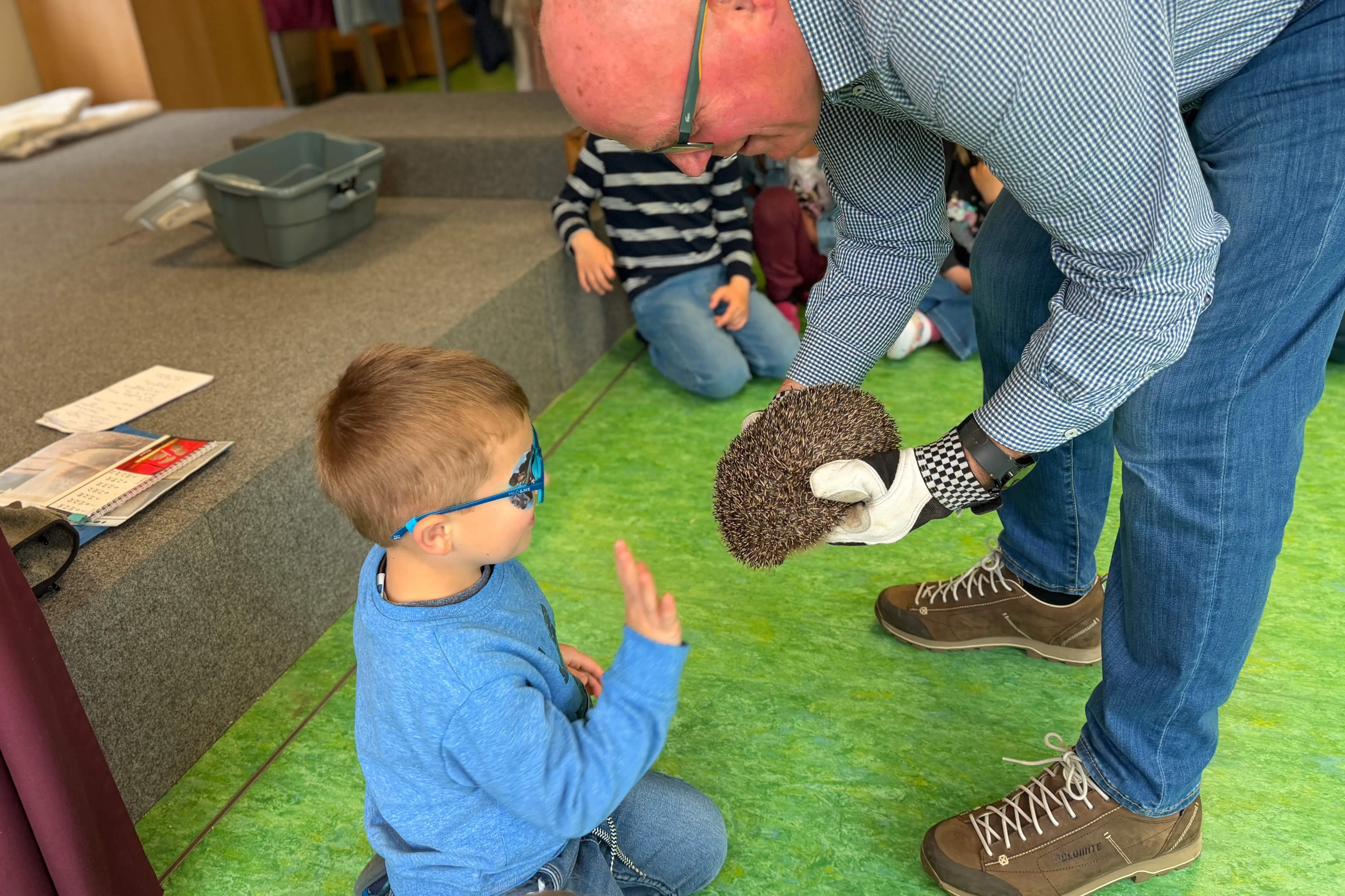 Ein Mann zeigt einem kleinen Jungen einen Igel, während andere Kinder im Hintergrund sitzen. Der Junge trägt eine Brille und streckt seine Hand aus.