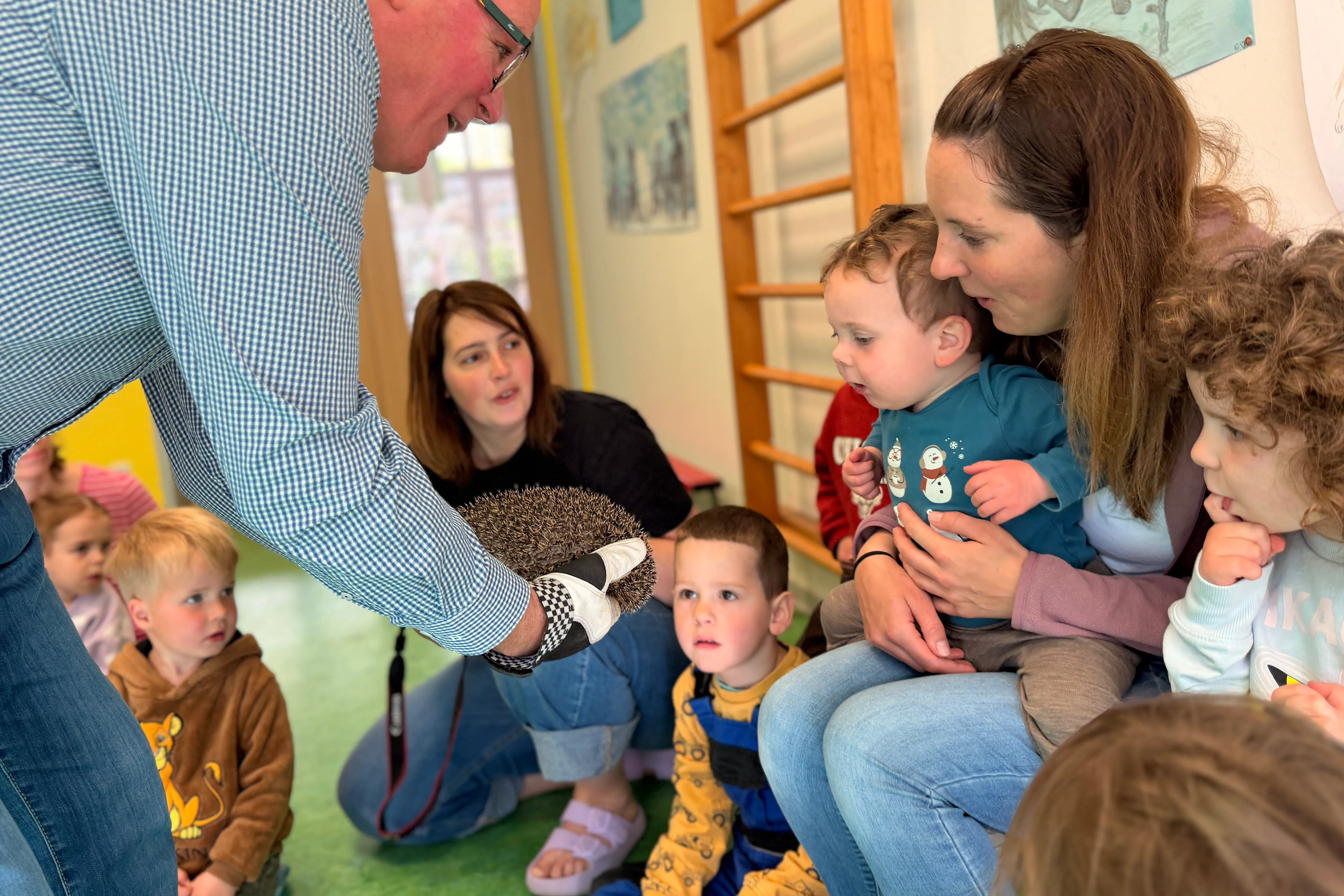 Ein Mann zeigt einen Igel einer Gruppe neugieriger Kinder in einem Kindergartenraum. Eine Frau hält ein Baby auf dem Arm und beobachtet das Geschehen.