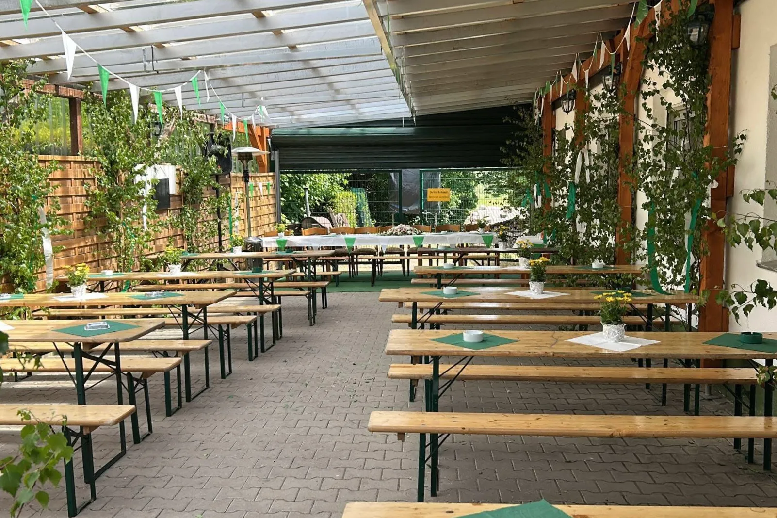Biergartentische und -bänke unter einem überdachten Bereich vor einem Gebäude mit Holzfassade und Pflanzenbewuchs.