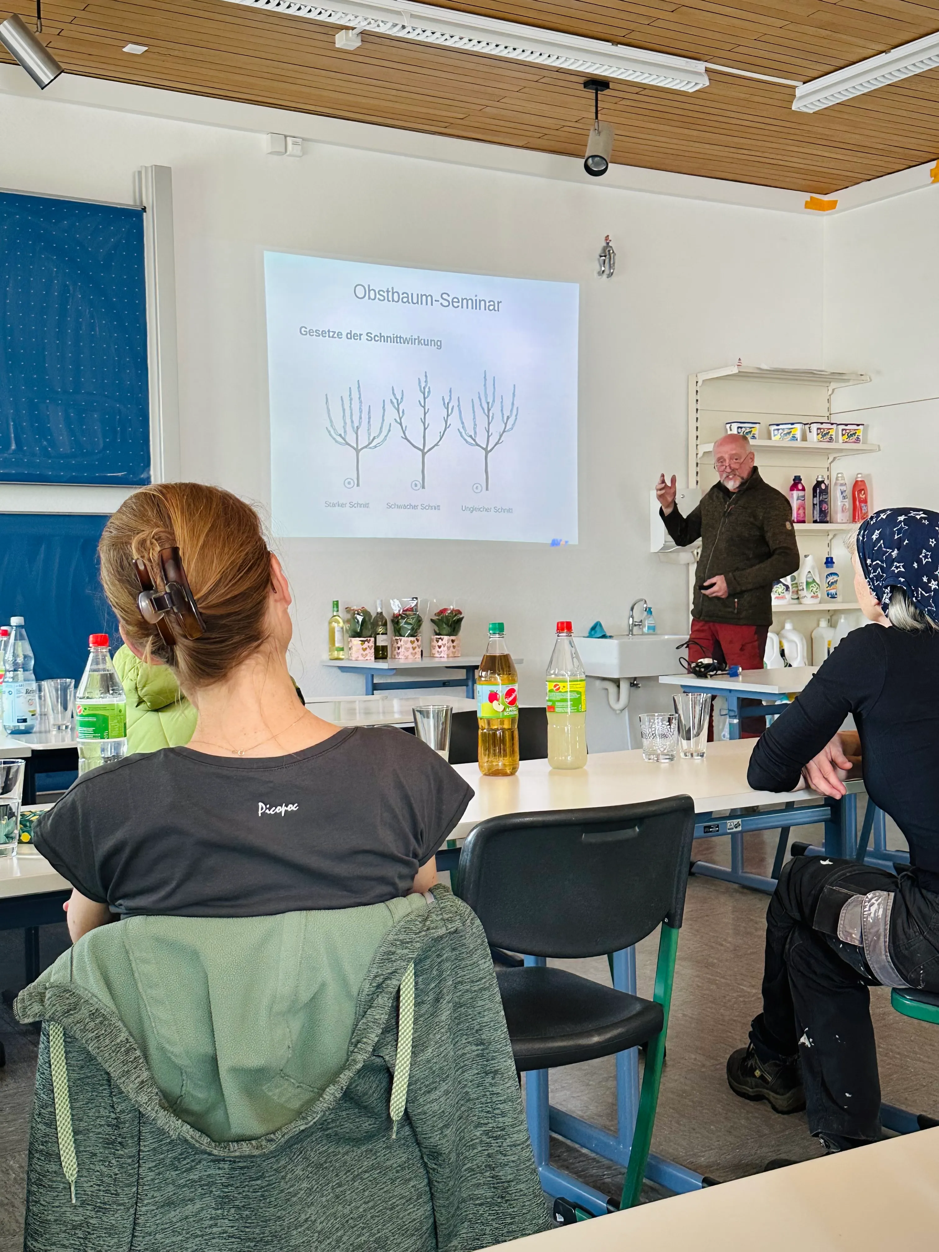 Ein Mann steht vor einer Projektion mit Diagrammen zum Thema Obstbaumschnitt und erklärt den Teilnehmern eines Seminars etwas. Im Vordergrund sitzen Teilnehmer, einige mit Notizen.