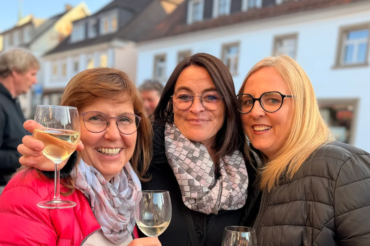 Drei lächelnde Frauen mit Weingläsern vor Häusern in Breckerfeld, im Hintergrund ein Mann. Der Feierabend markiert das vierjährige Bestehen des Vereins LokalLaden.