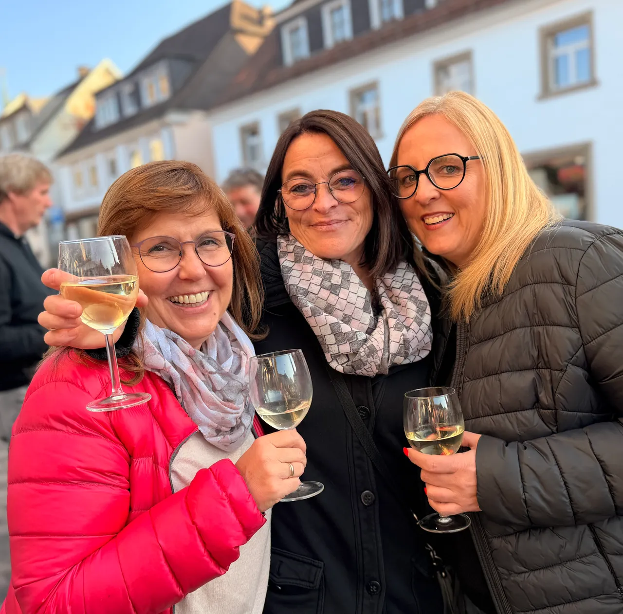Drei lächelnde Frauen mit Weingläsern posieren eng umschlungen vor einer hellen Fassade. Im Hintergrund sind weitere Menschen zu erkennen.