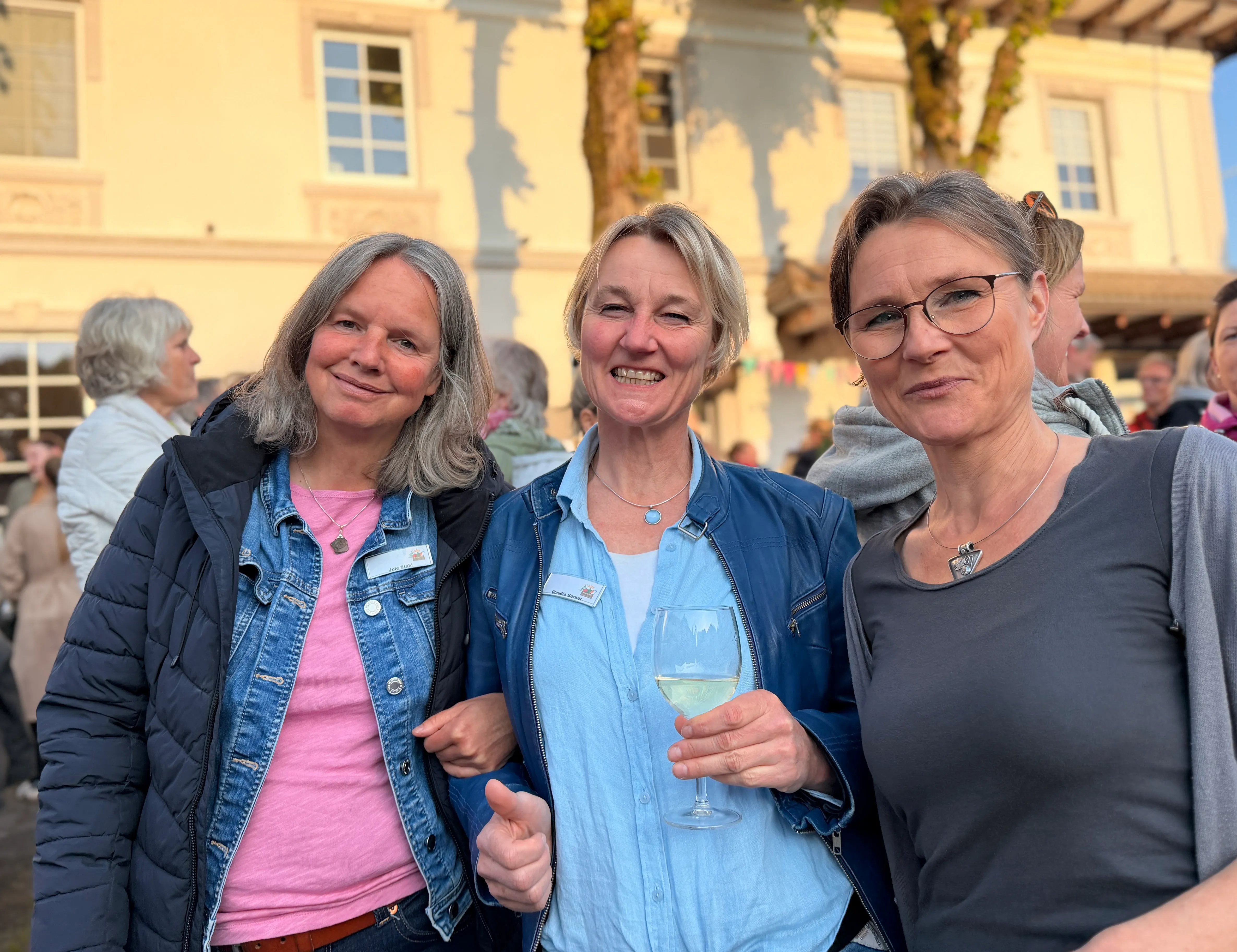 Drei Frauen lächeln in die Kamera, die mittlere hält ein Weinglas. Im Hintergrund ein helles Gebäude und weitere Menschen.