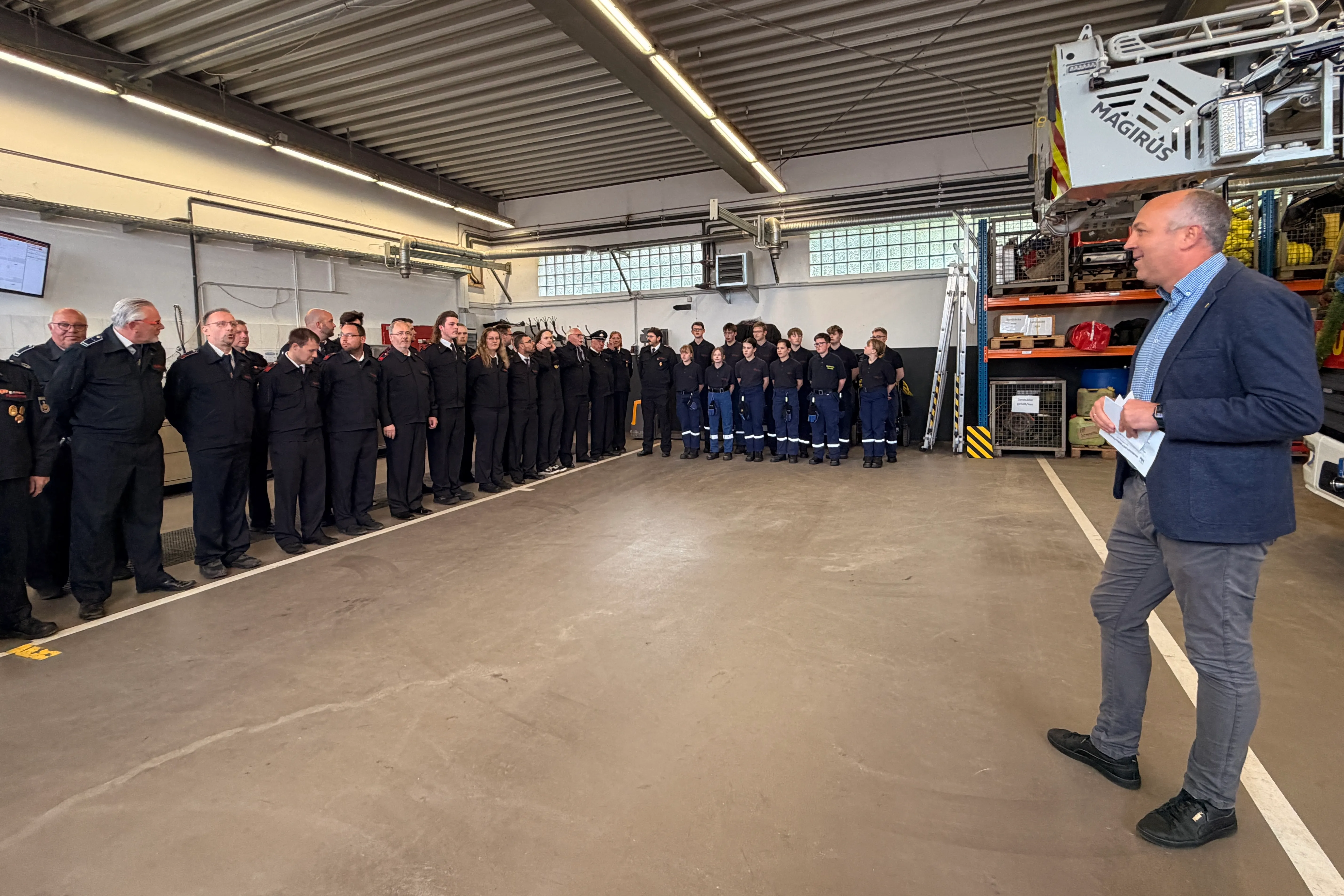 Ein Mann in einem Blazer hält Papiere und spricht vor einer Reihe von Feuerwehrleuten in Uniform in einer großen Werkstatthalle.