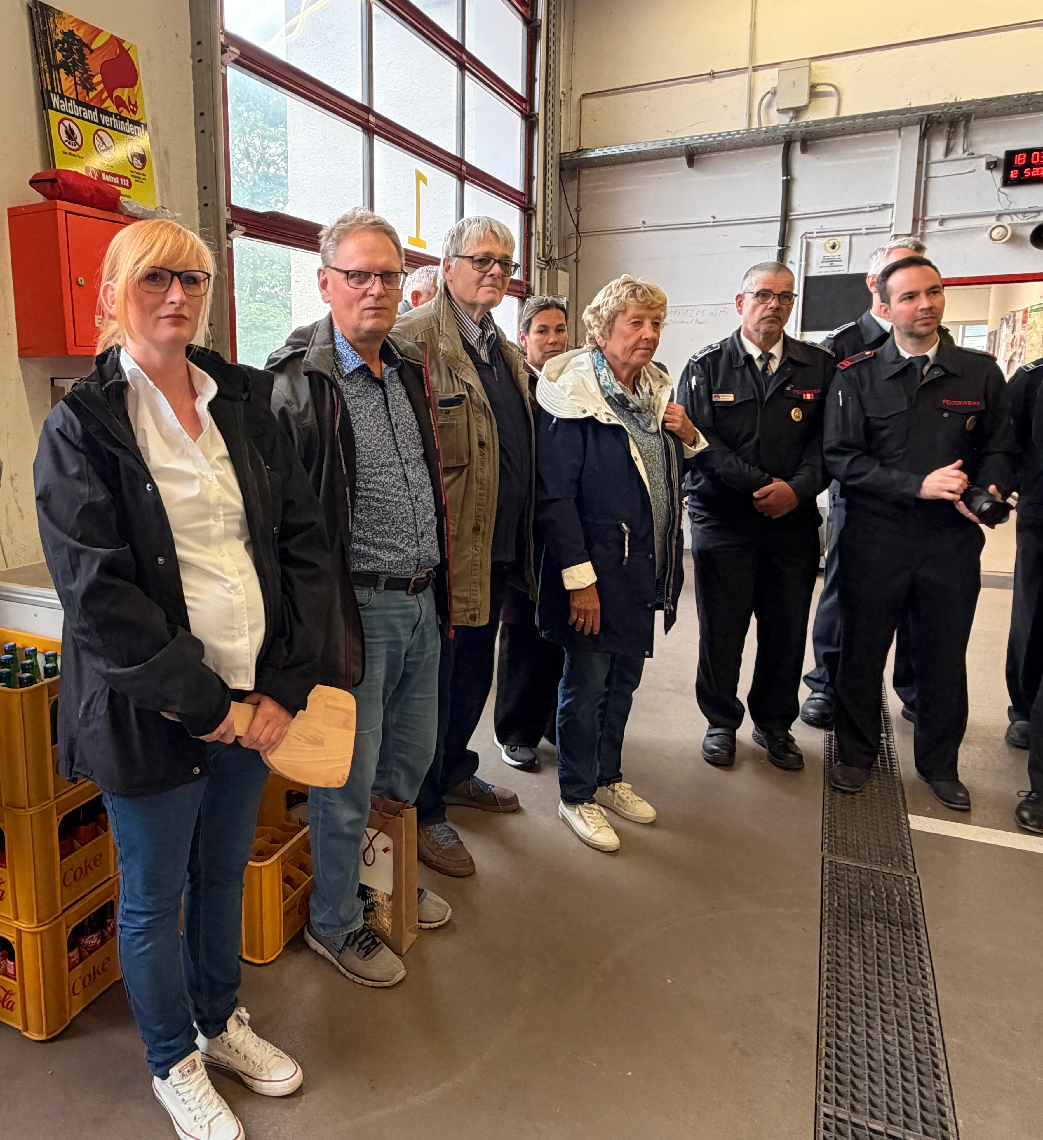 Gruppe von Menschen, darunter Feuerwehrleute in Uniform und Zivilpersonen, steht vor einer geschlossenen Garagentür und blickt in eine Richtung.