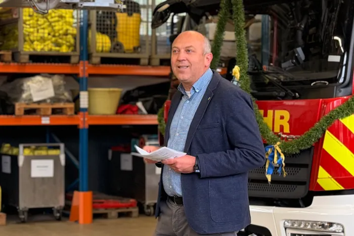 Ein Mann in einem Blazer hält Papiere vor einem roten Feuerwehrauto in einer Lagerhalle. Im Hintergrund Regale mit gelben Kisten.