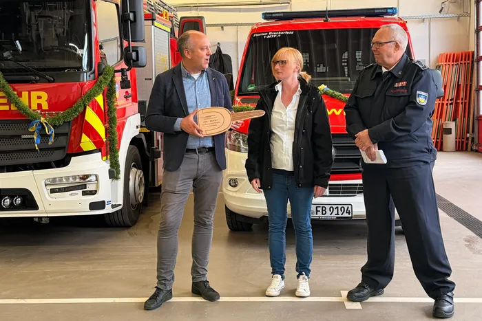 Bürgermeister André Dahlhaus überreicht Wehrleiter Andreas Bleck einen symbolischen Schlüssel, während Miriam Link von Magirus daneben steht. Im Hintergrund ein rotes Feuerwehrfahrzeug.