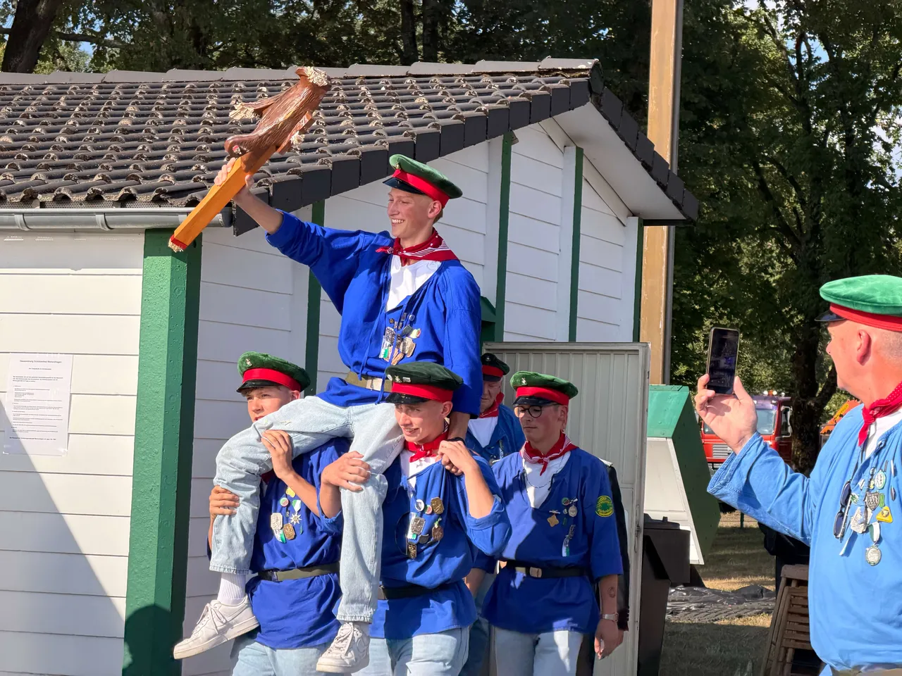 Ein Mann in traditioneller Kleidung mit grüner Mütze wird von mehreren Personen auf den Schultern getragen und hält einen hölzernen Gegenstand hoch. Im Hintergrund steht ein Gebäude und weitere Personen in ähnlicher Kleidung sind zu sehen.
