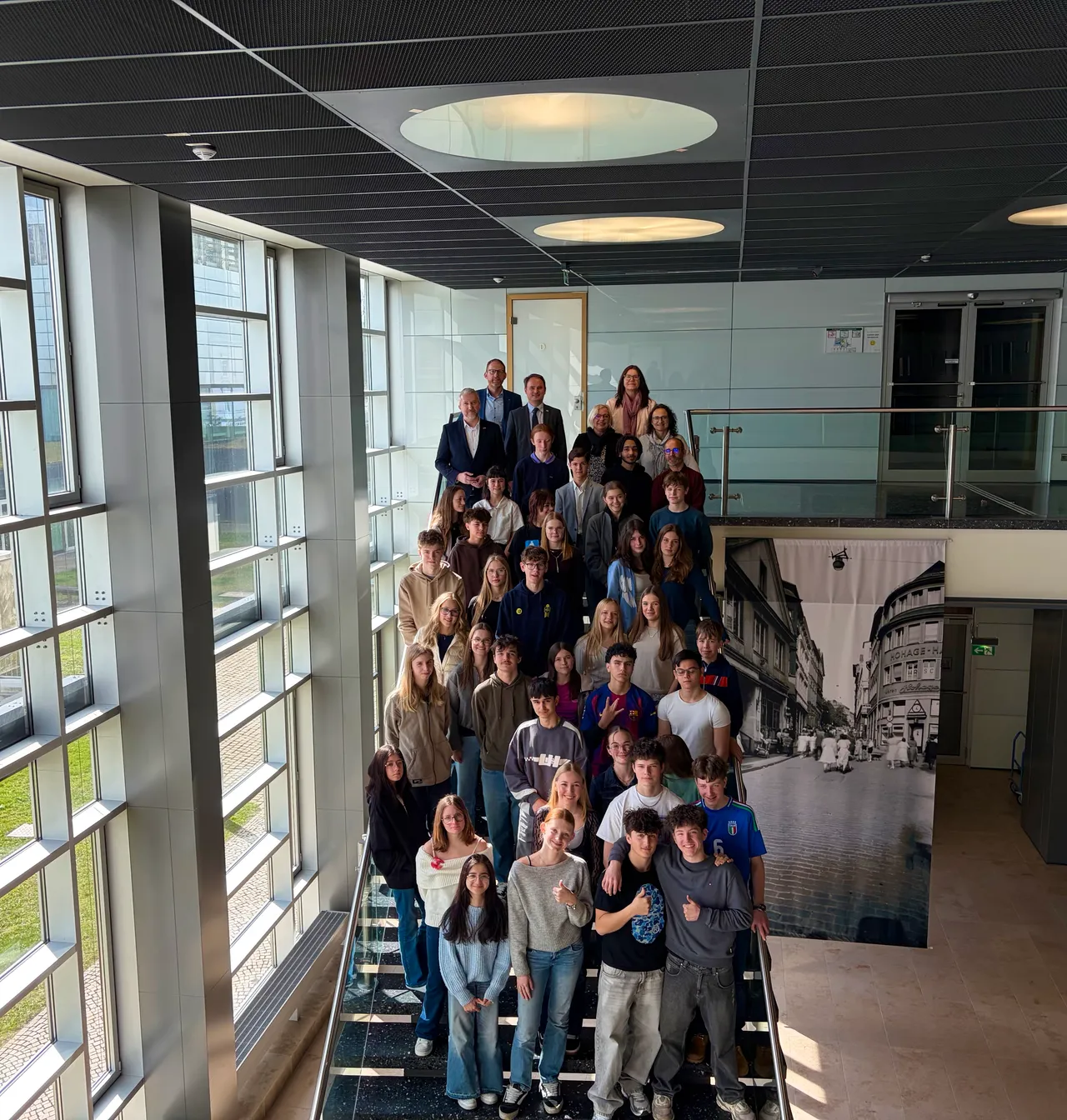 Eine Gruppe von etwa 30 Jugendlichen und einigen Erwachsenen posiert auf einer Treppe in einem modernen Gebäude, im Hintergrund ein großes historisches Stadtbild.