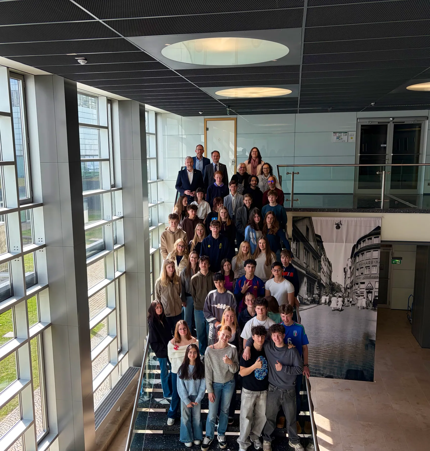 Eine Gruppe von etwa 30 Jugendlichen und einigen Erwachsenen posiert auf einer Treppe in einem modernen Gebäude, im Hintergrund ein großes historisches Stadtbild.