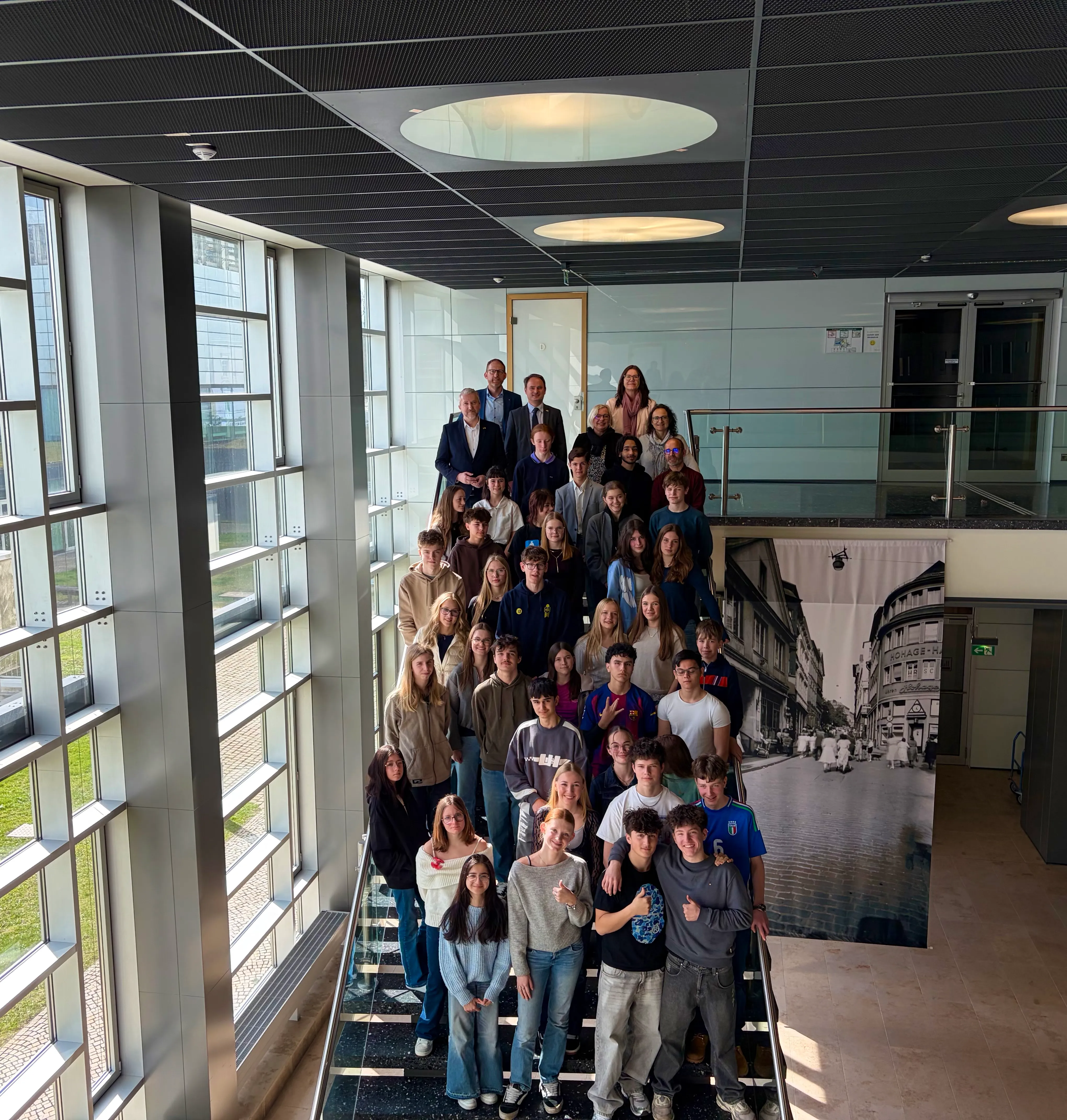 Eine Gruppe von etwa 30 Jugendlichen und ein Mann posieren auf einer Treppe in einem modernen Gebäude mit Glasfassade. Im Hintergrund ein großes Foto einer historischen Stadtansicht.