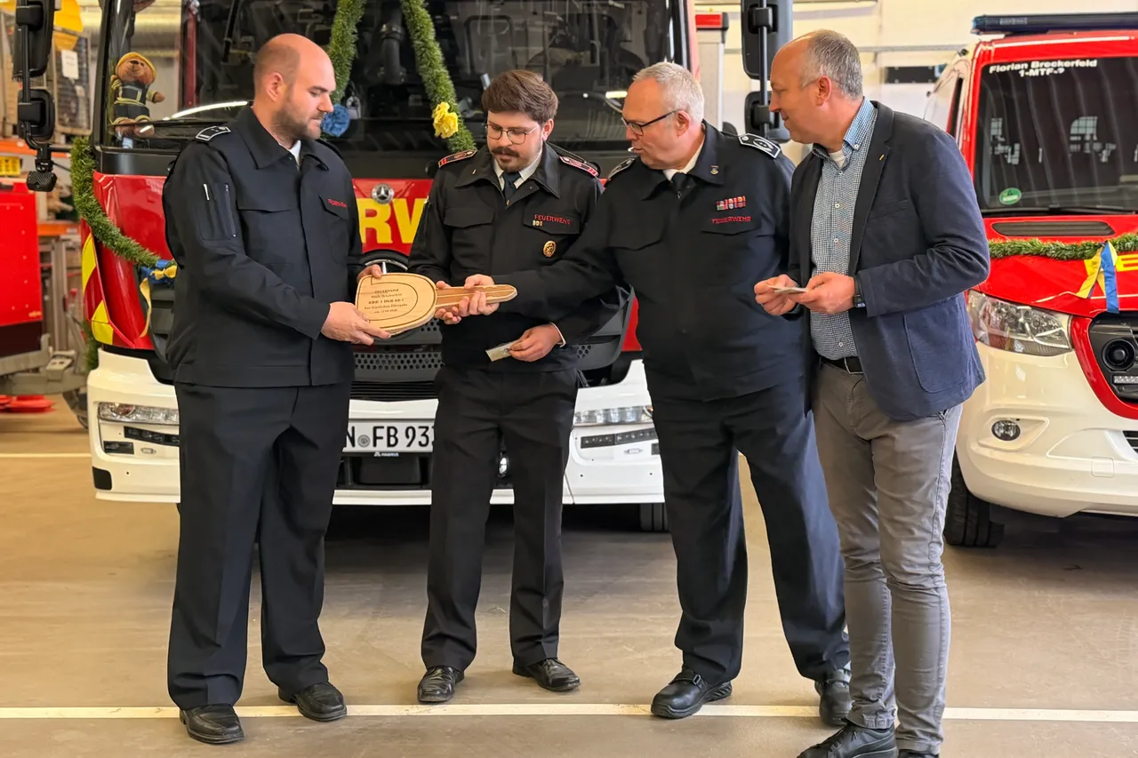 Vier Männer stehen vor einem weißen Mannschaftswagen der Feuerwehr und betrachten ein Geschenk – eine gravierte Holztafel. Im Hintergrund sind ein rotes Feuerwehrauto und ein weiterer Wagen zu sehen.