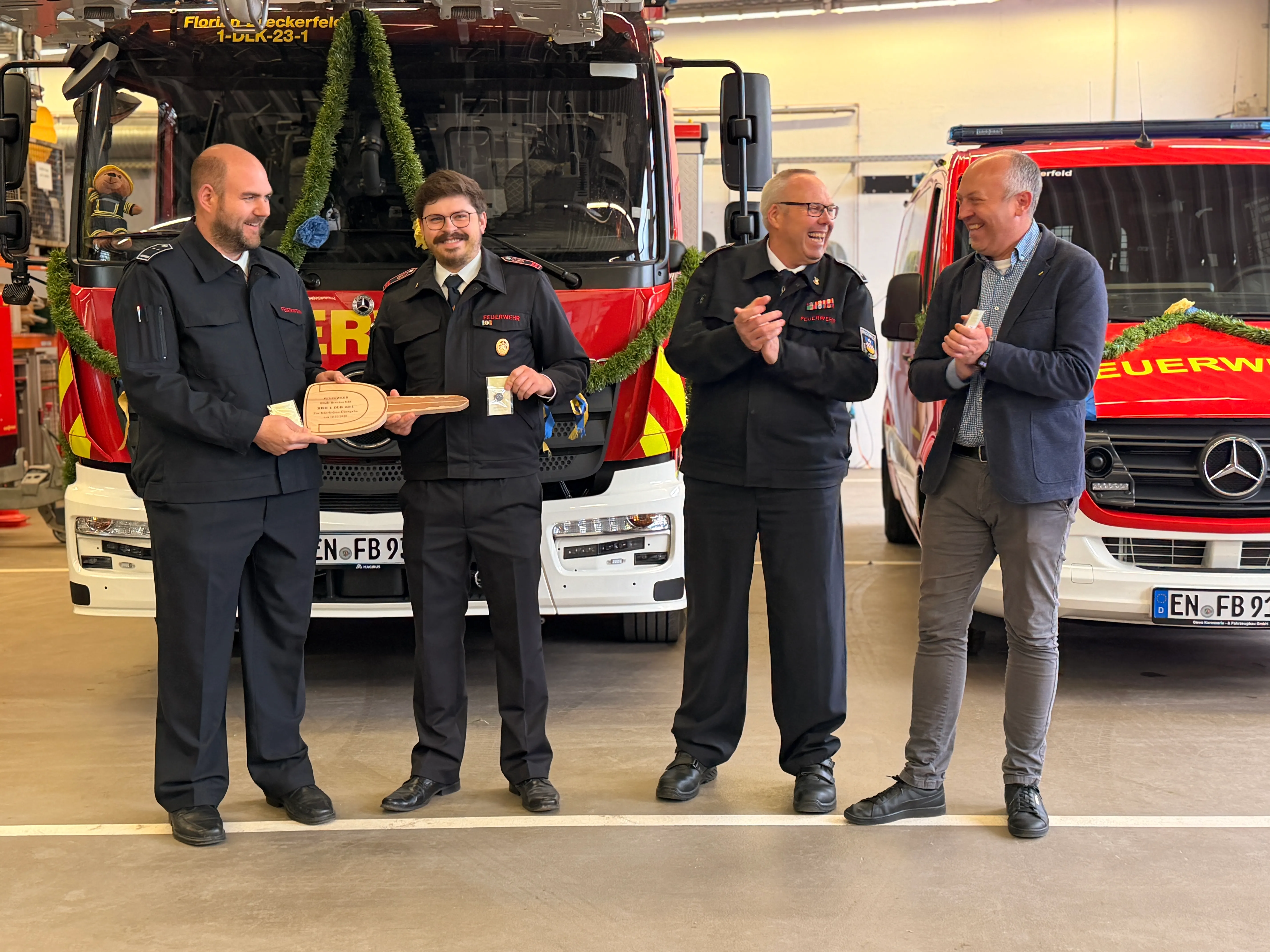 Vier Männer stehen vor einem neuen roten Feuerwehrauto und applaudieren. Zwei Feuerwehrleute in Uniform halten eine hölzerne Skulptur entgegen.
