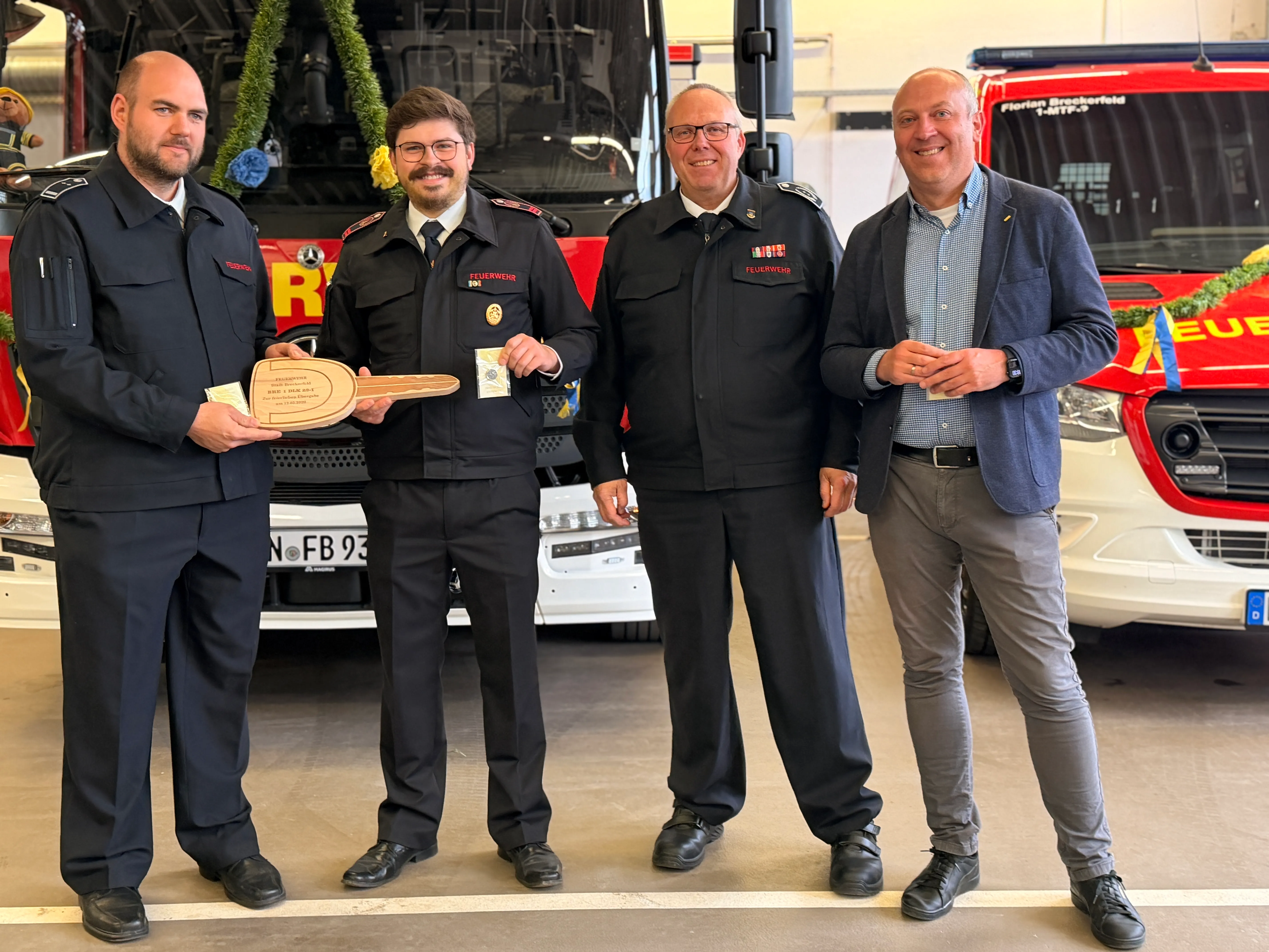 Vier Männer stehen vor einem roten Feuerwehrauto. Zwei von ihnen tragen Uniformen der Freiwilligen Feuerwehr, die anderen zwei sind zivil gekleidet und halten Geschenke.