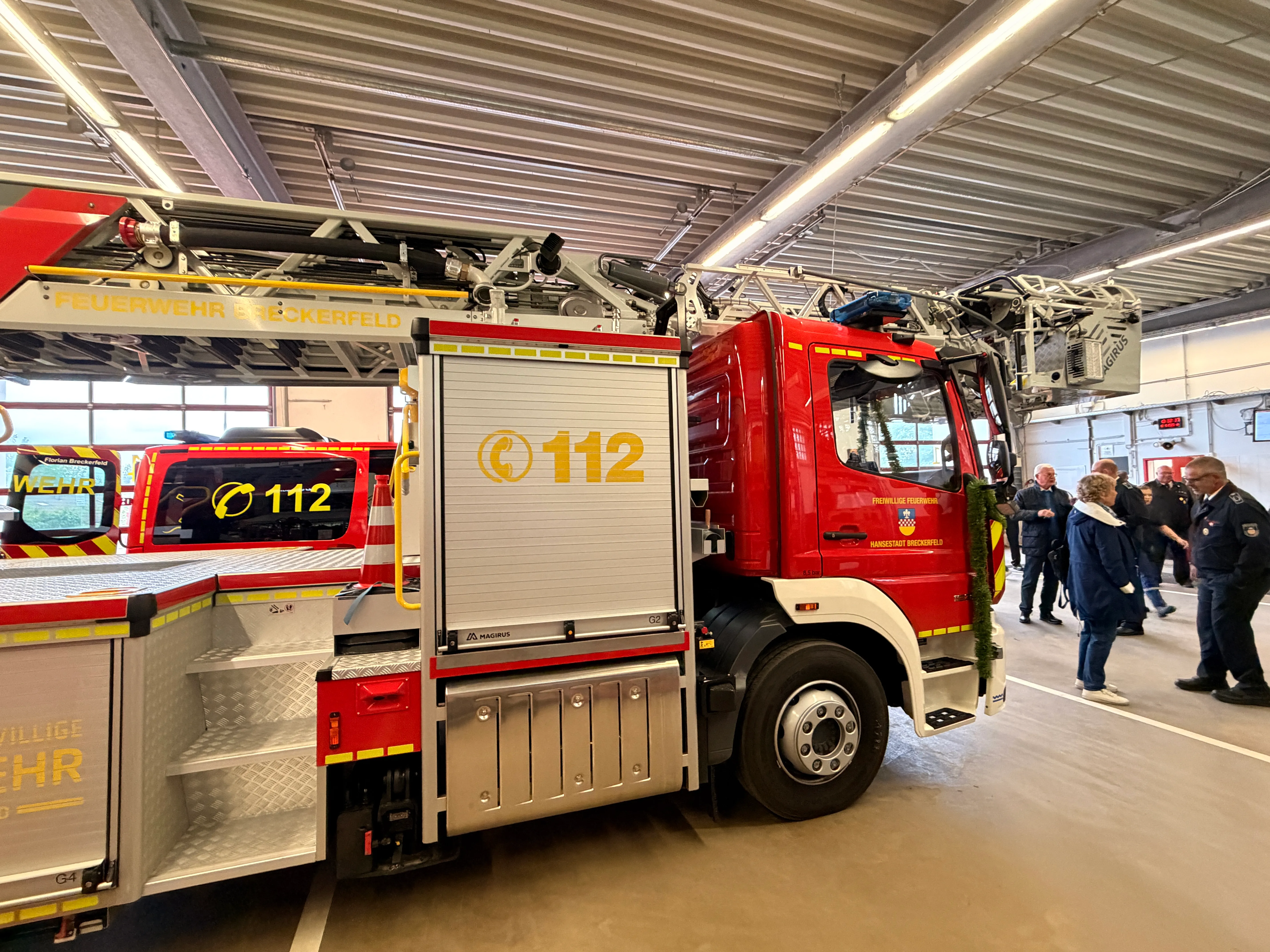 Roter Feuerwehrauto mit der Aufschrift „Feuerwehr Breckerfeld“ und der Notrufnummer 112 steht in einer modernen Fahrzeughalle. Im Hintergrund sind weitere Einsatzfahrzeuge und Personen zu sehen.